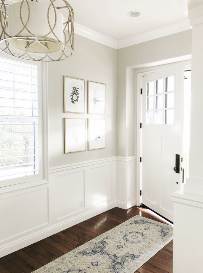 Bright entryway with white walls, a wooden floor, a patterned rug, a window with white shutters, and a closed white door with window panels.