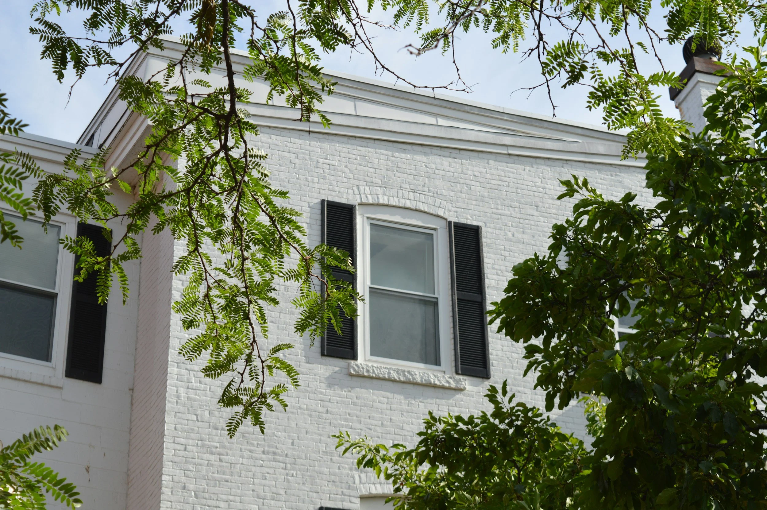 A white brick house with black window shutters, surrounded by green trees and leaves.