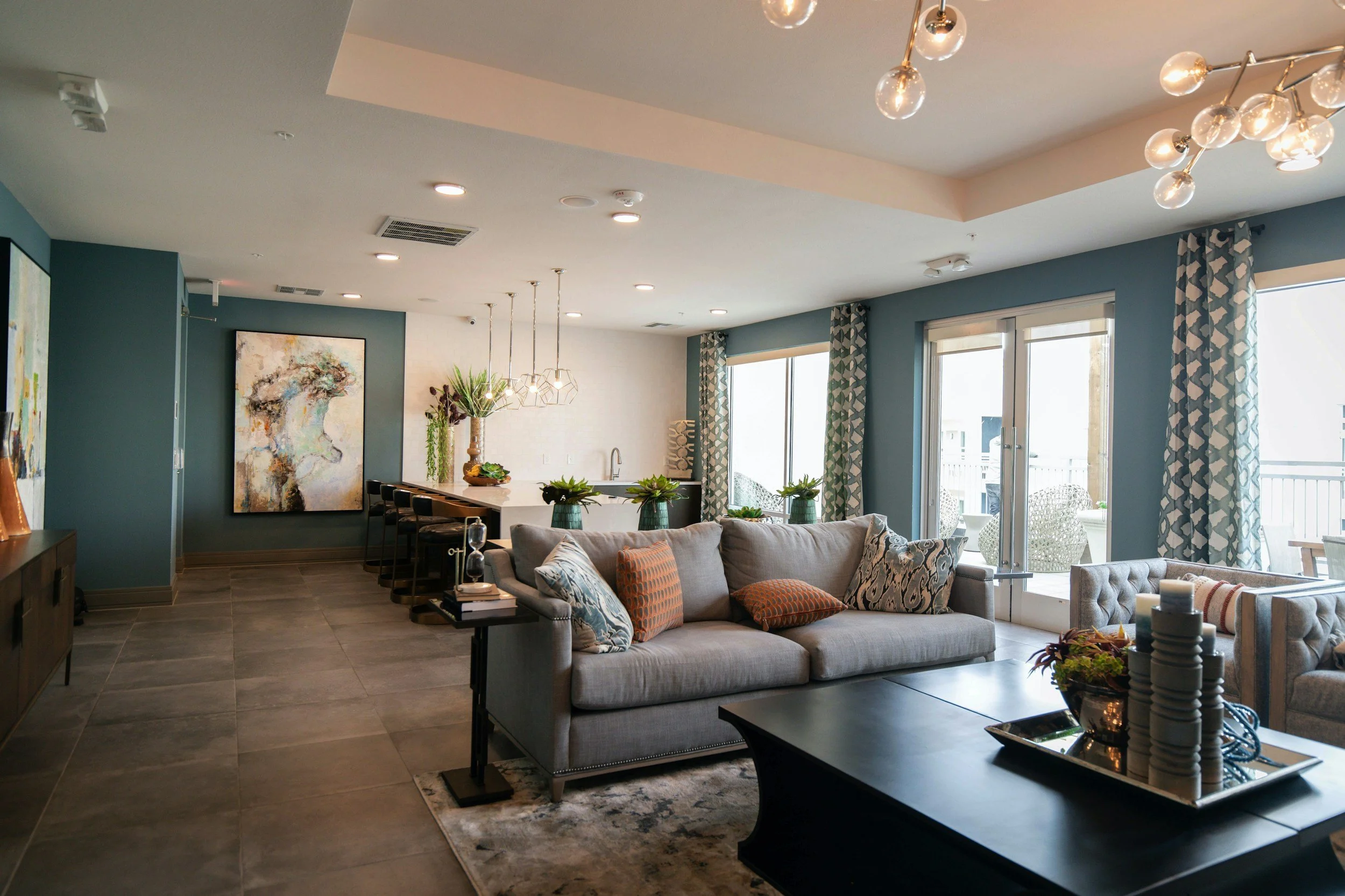 Open-concept living room and kitchen with blue walls, large windows, and modern decor including a sofa, armchair, coffee table with decorative items, and a kitchen island with barstools and pendant lighting.