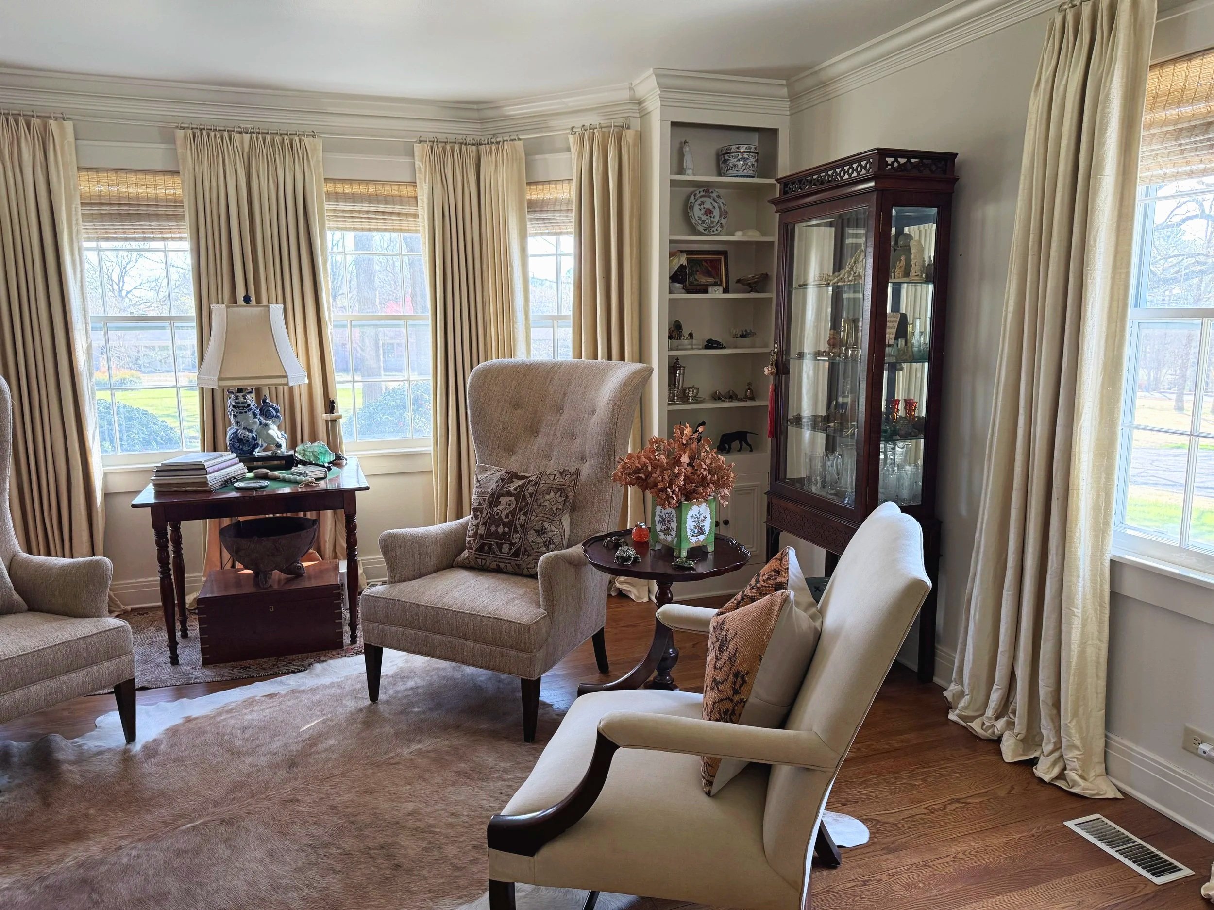 A well-decorated living room with beige armchairs, a side table with a vase of dried flowers, a glass display cabinet, and large windows with beige curtains showing a sunny outdoor view.