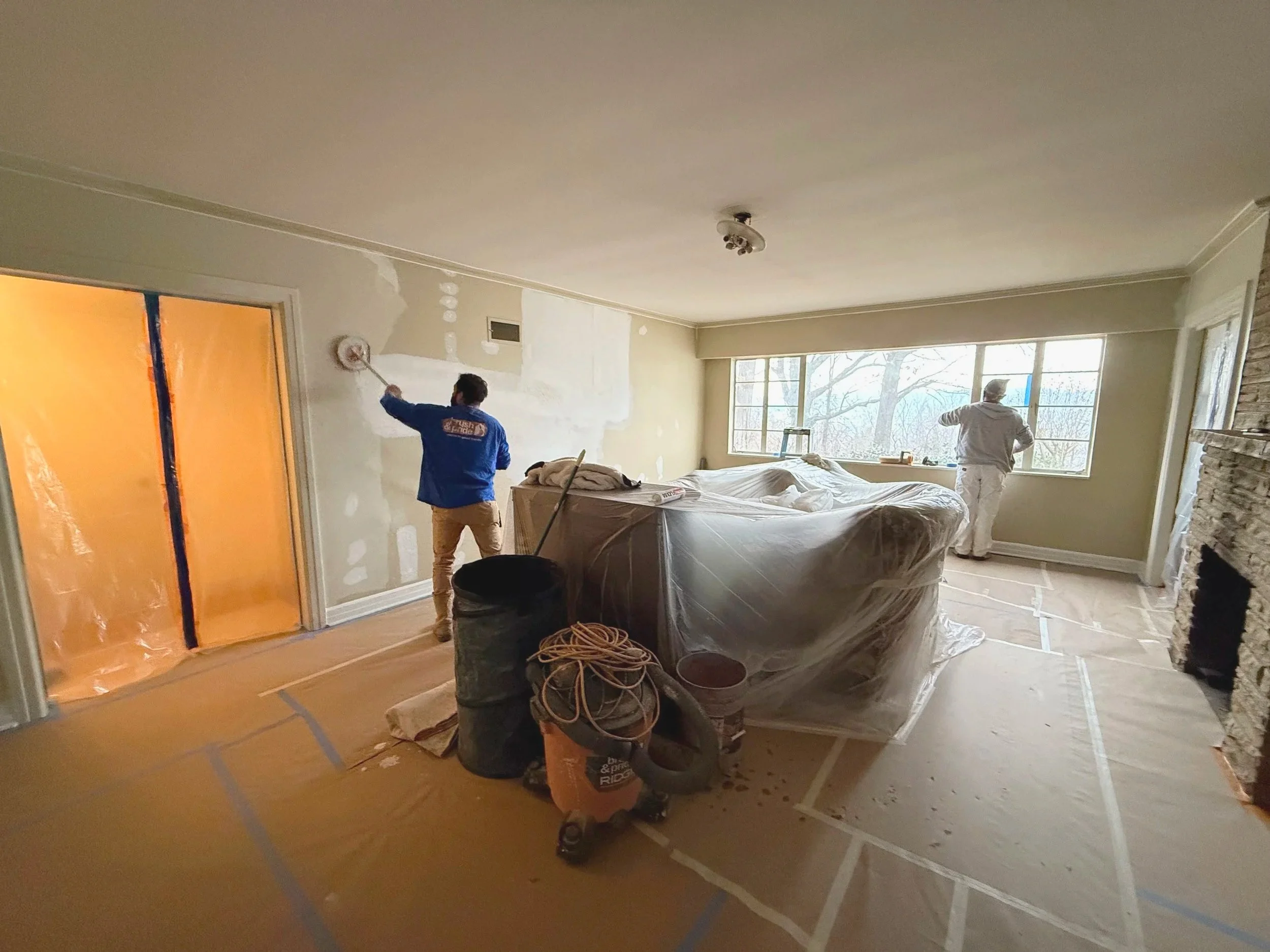 Two workers painting a living room wall, one near a sliding door and the other near a window, with furniture and construction supplies in the room.