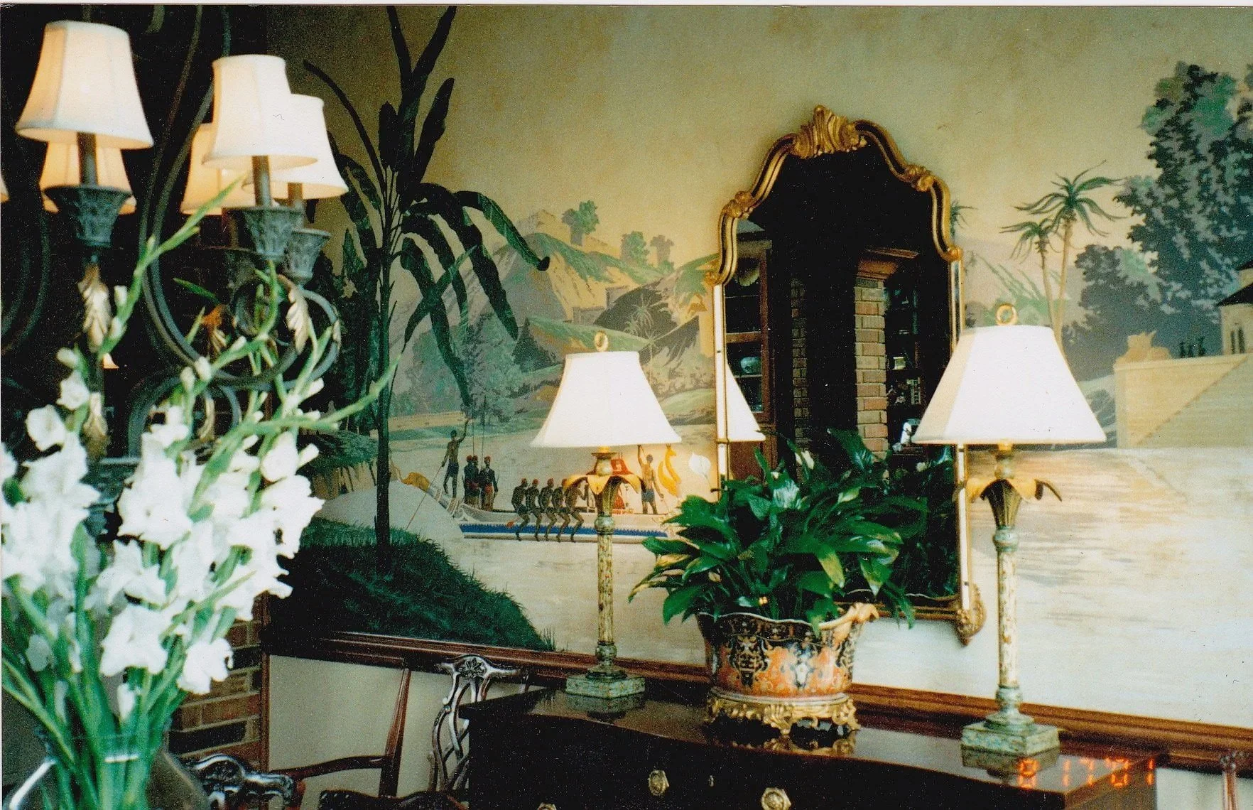 Interior of a room with a lamp on a wooden cabinet, a framed mirror, a potted plant, and a detailed painted mural featuring a landscape and people in a boat.