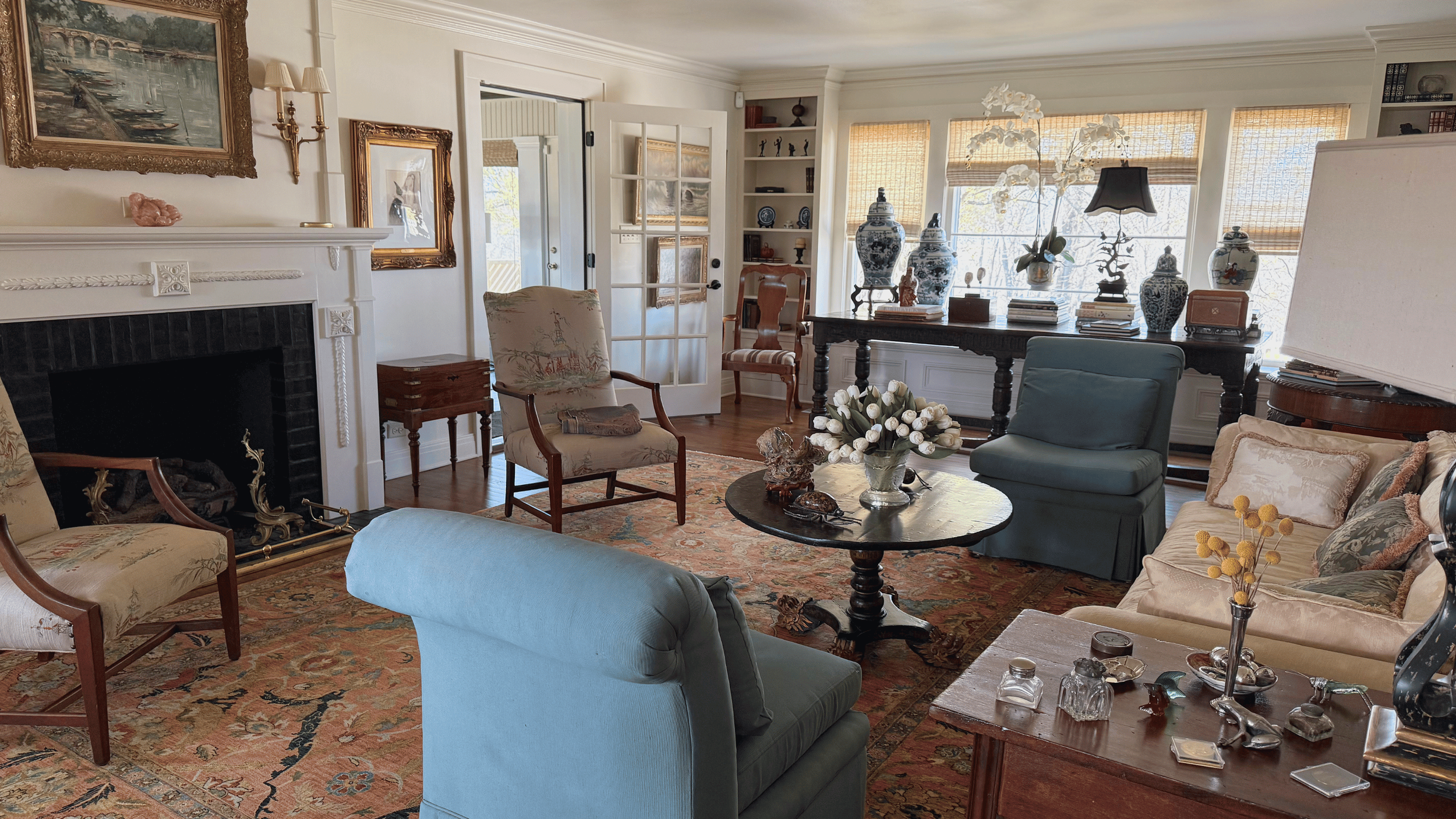 A cozy living room with a fireplace, multiple upholstered chairs, a large dark wood table with decorative vases, books, and ornaments, and large windows with bamboo shades allowing sunlight inside.