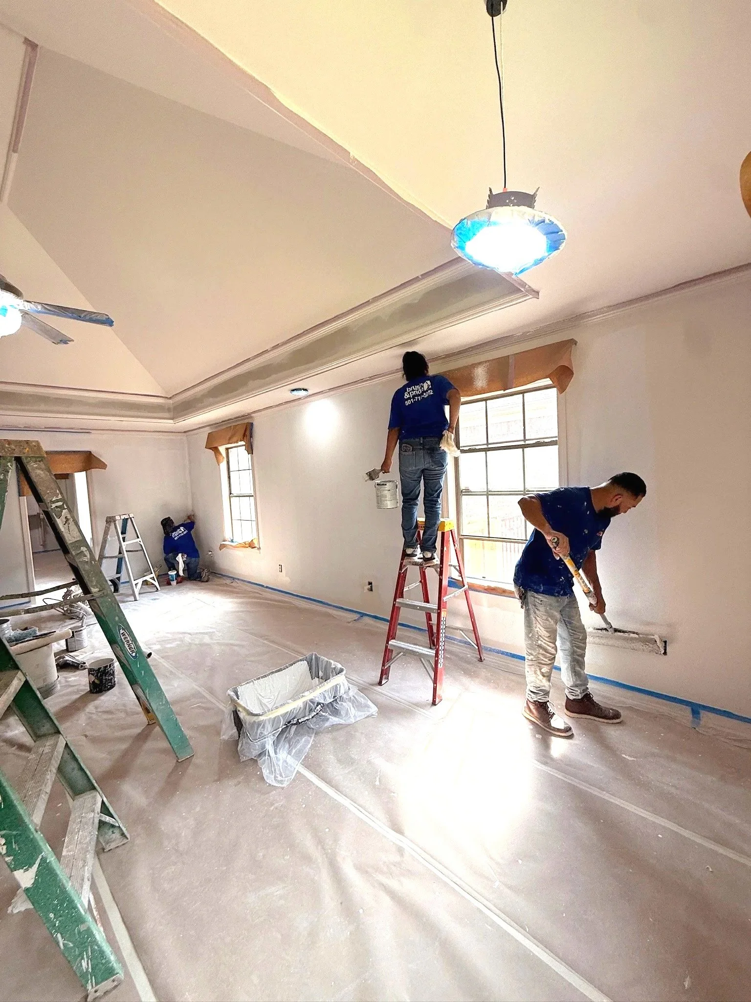Workers painting walls and ceilings in a room with large windows and various ladders, buckets, and tools scattered around.
