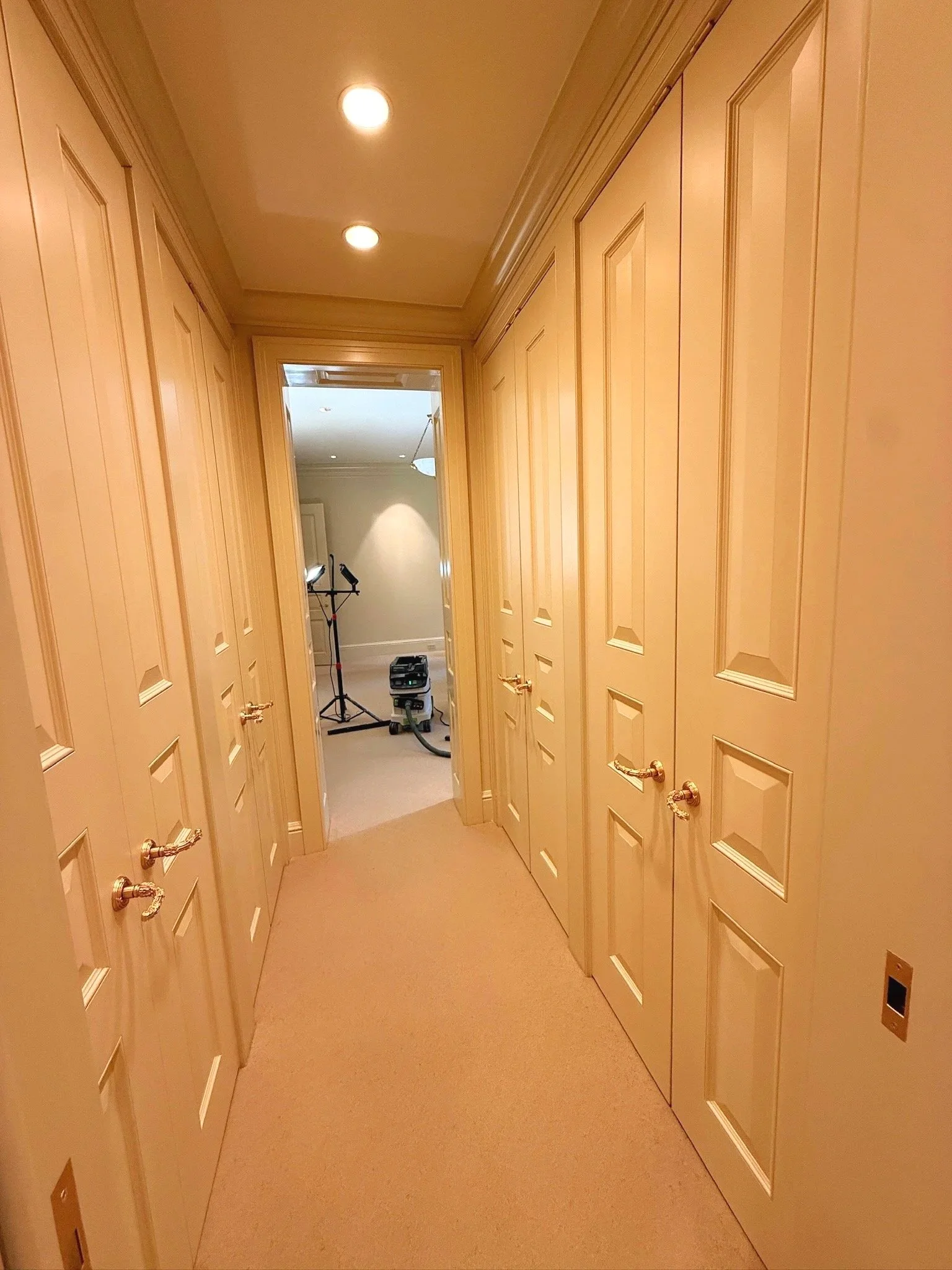A hallway with beige carpet and wooden-paneled closet doors on both sides, leading to a room with a stand with equipment and a lamp in the background.