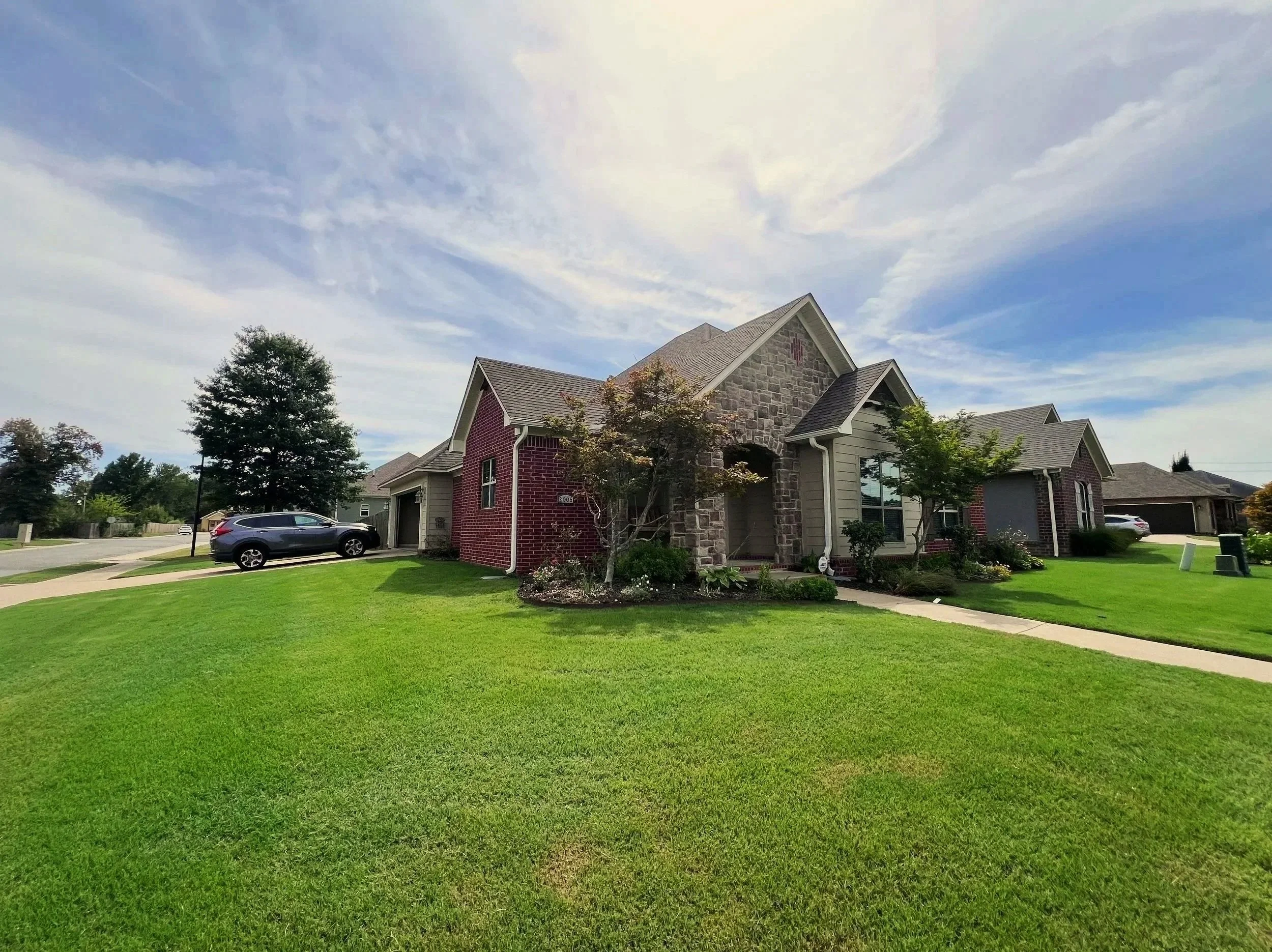 A suburban house with stone and brick exterior, front yard with green grass, small trees, sidewalk, and parked cars on a driveway under a blue sky with scattered clouds.