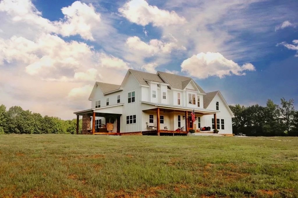 Large white house with multiple stories and a spacious porch, situated on a grassy field with trees in the background and a partly cloudy sky overhead.