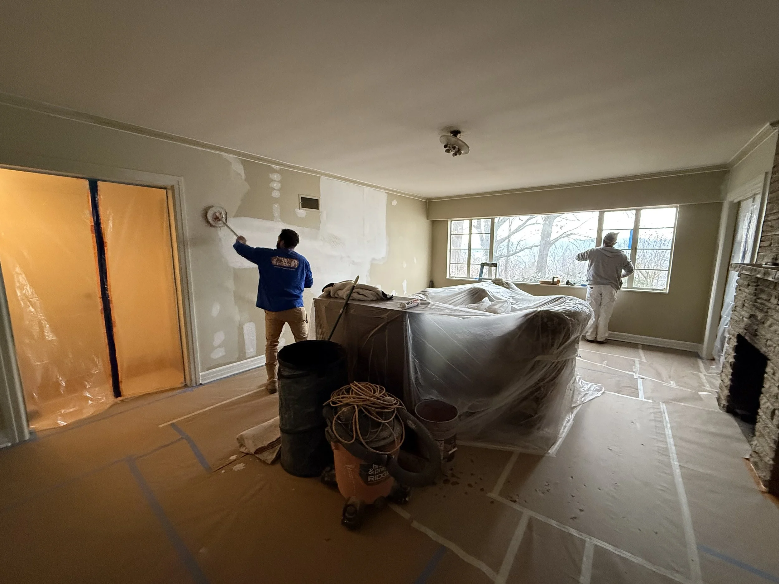 Two workers painting a living room wall, one near a sliding door and the other near a window, with furniture and construction supplies in the room.
