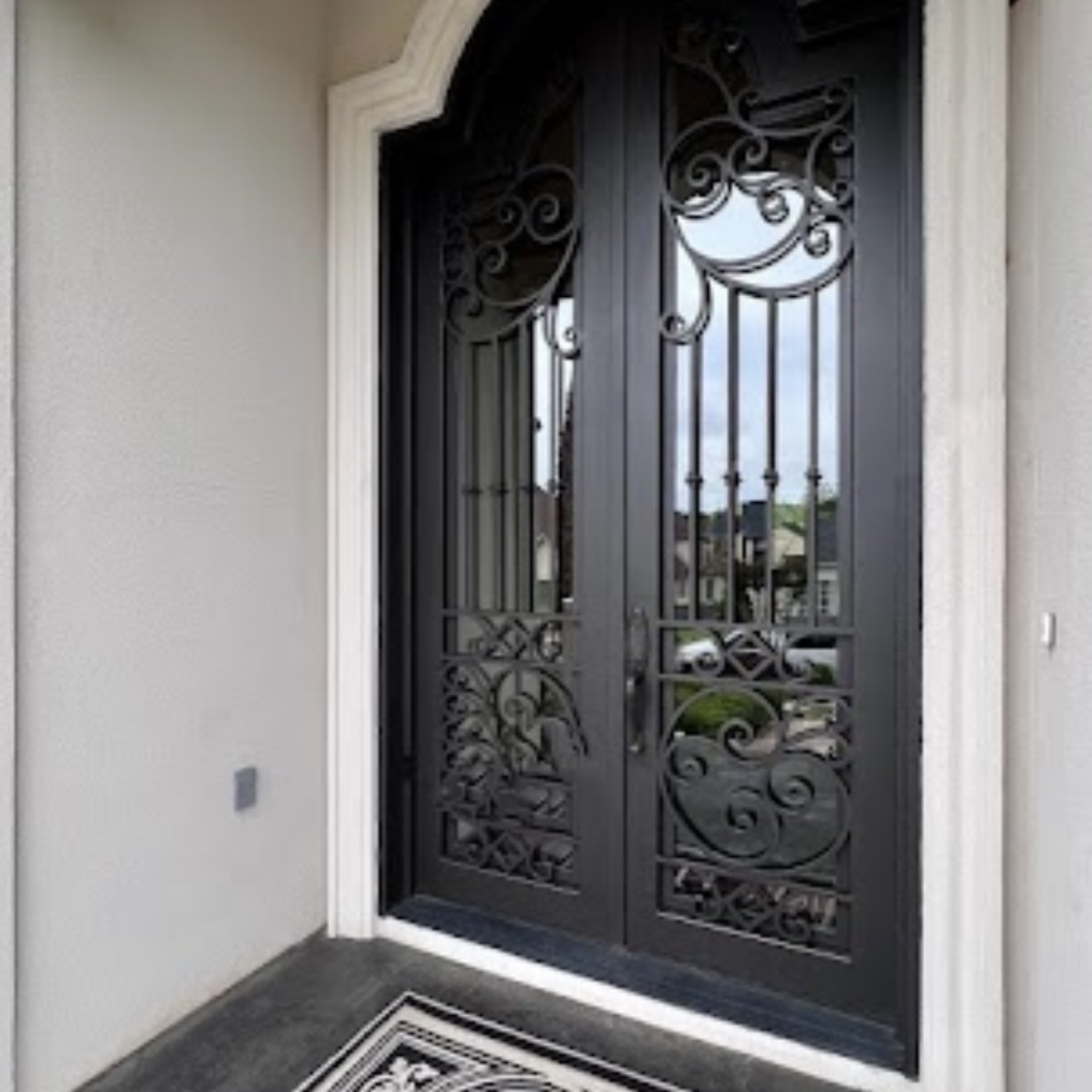 Decorative black metal front door with intricate swirl patterns, framed by light-colored walls, with a black and white outdoor rug in front.