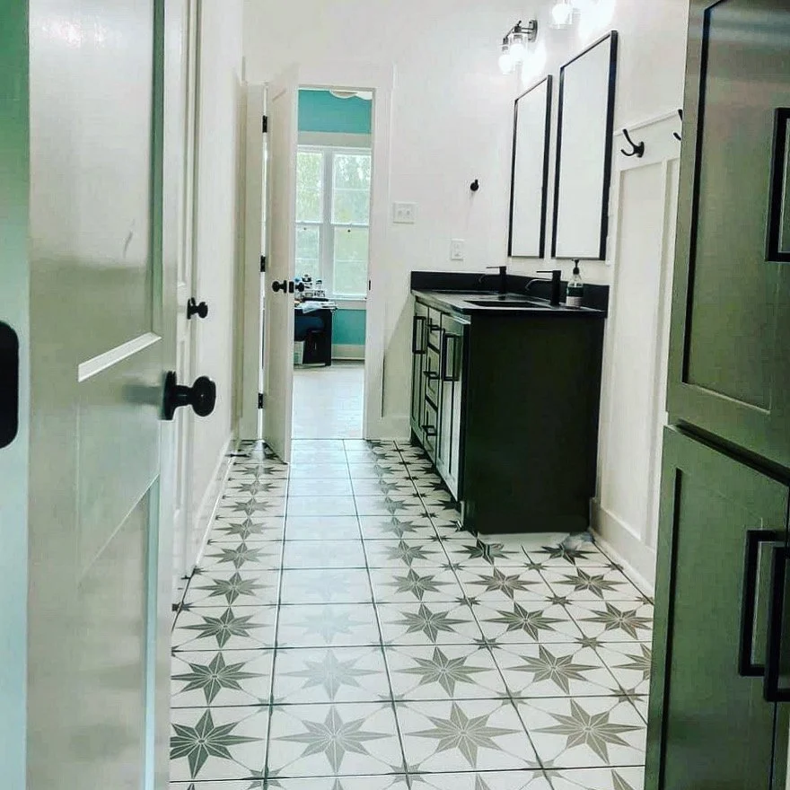 A narrow kitchen with white walls and patterned tile floor, featuring black and white cabinets, a black countertop, a double sink, and two framed mirrors above the sink. There is a doorway leading to a bright room with windows.