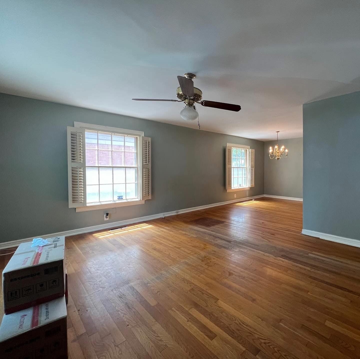Empty living room with hardwood floors, two windows with white shutters, a ceiling fan, and a small chandelier, with a box and some paper on the floor.