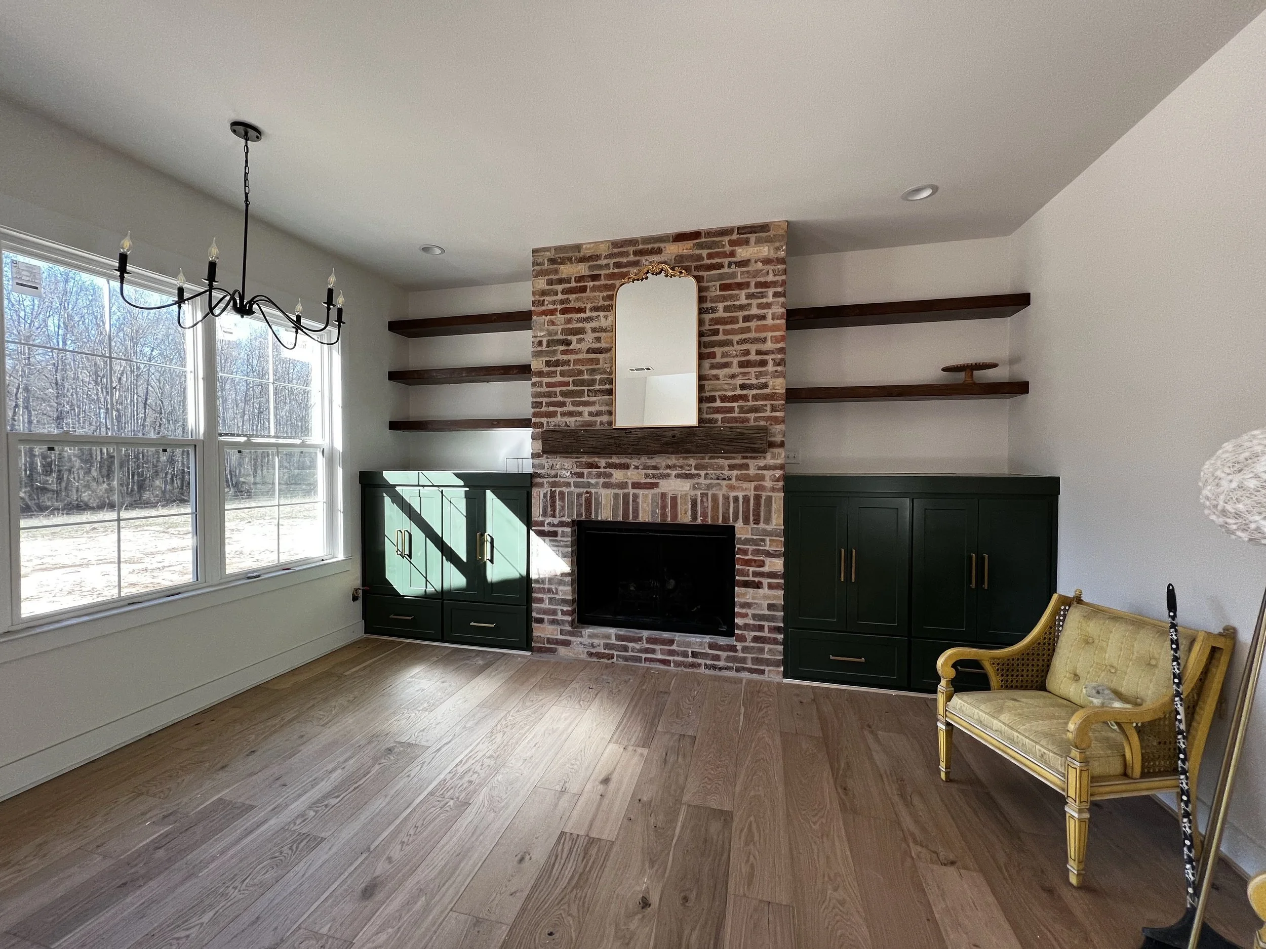 Living room with large windows, a brick fireplace with a mirror above, dark green cabinets, wooden shelves, a yellow vintage settee, and a black chandelier.