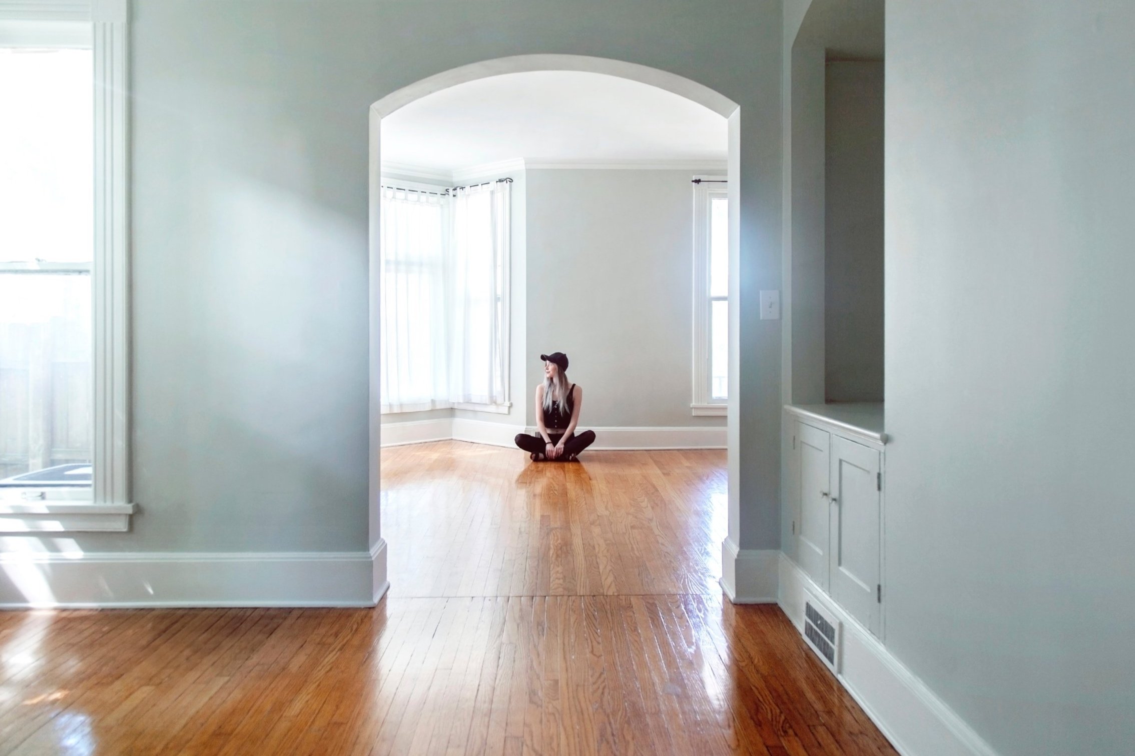 A person sitting cross-legged on wooden floor in a bright, empty living room with white walls and large windows with white curtains.