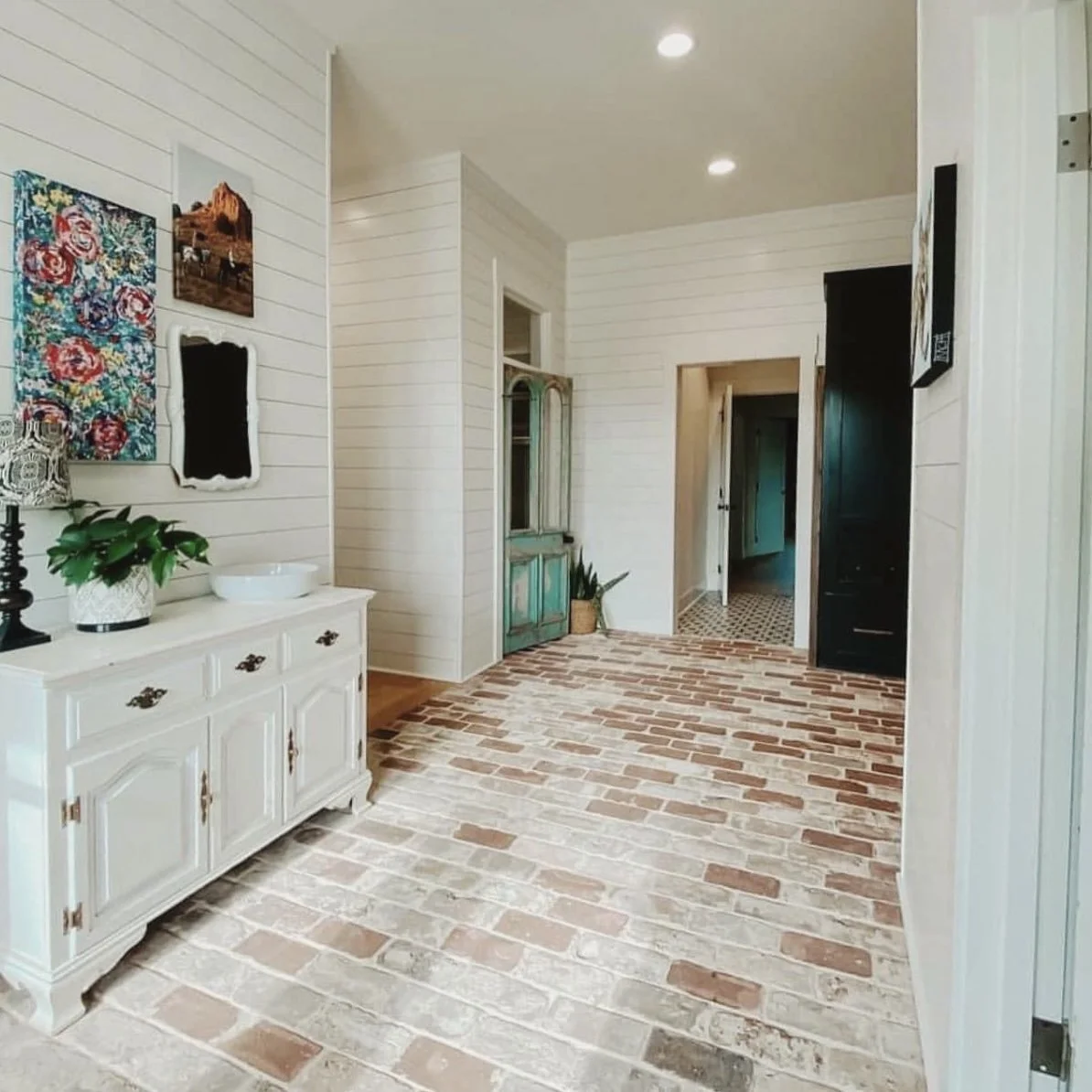 Entryway with white shiplap walls, brick flooring, a white sideboard with a potted plant and lamp, colorful artwork, a distressed blue cabinet, and hallway leading to other rooms.