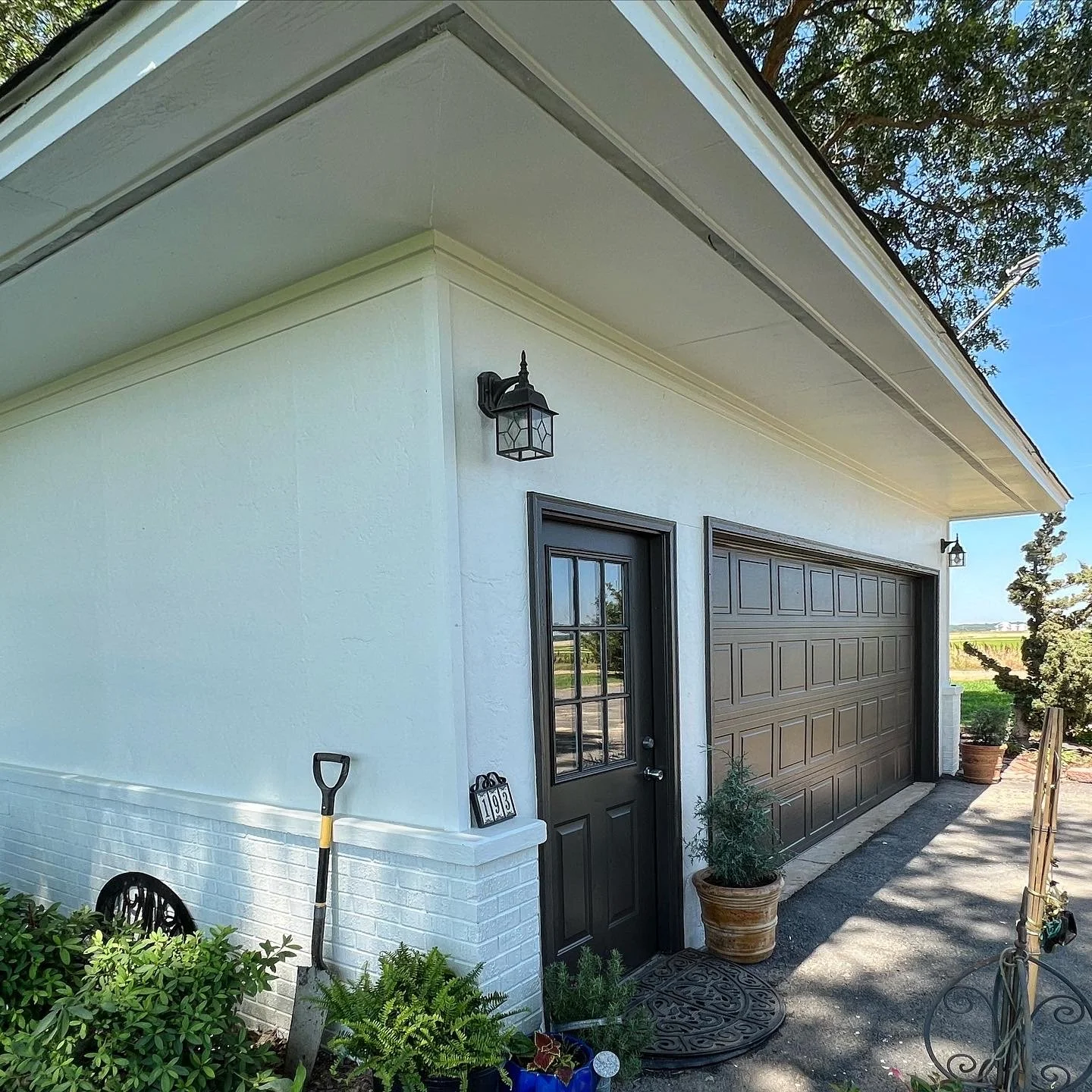 A white house garage with a black garage door and a black side door, outdoor lanterns, potted plants, a shovel, and a garden area during daytime.