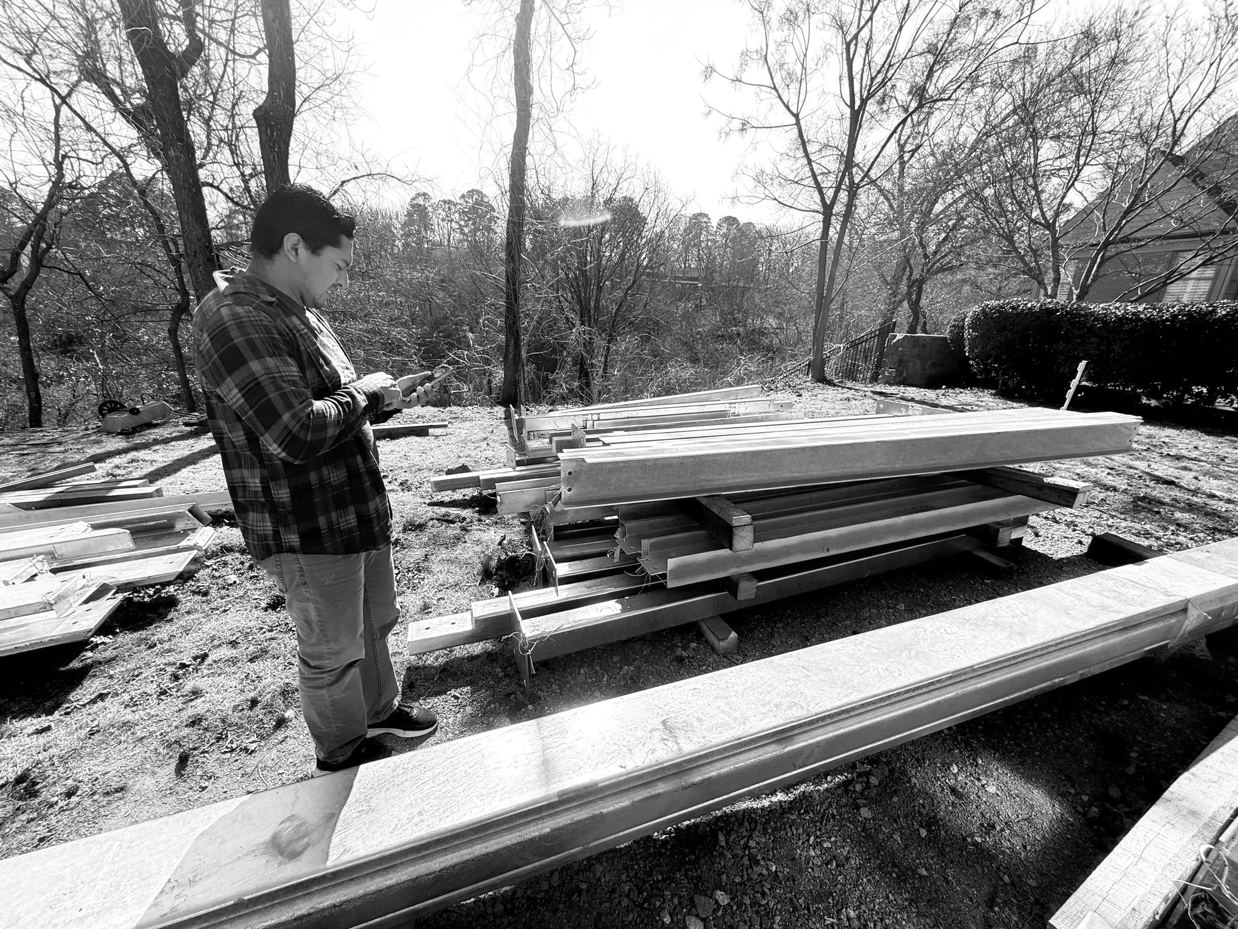 A man standing outdoors on a construction site, looking at his phone, with stacked lumber and trees in the background.