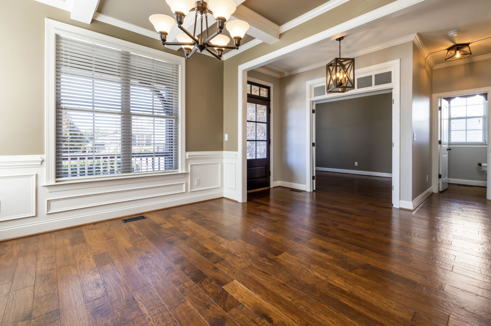 Empty living room with hardwood floors, large window with blinds, and modern light fixtures.