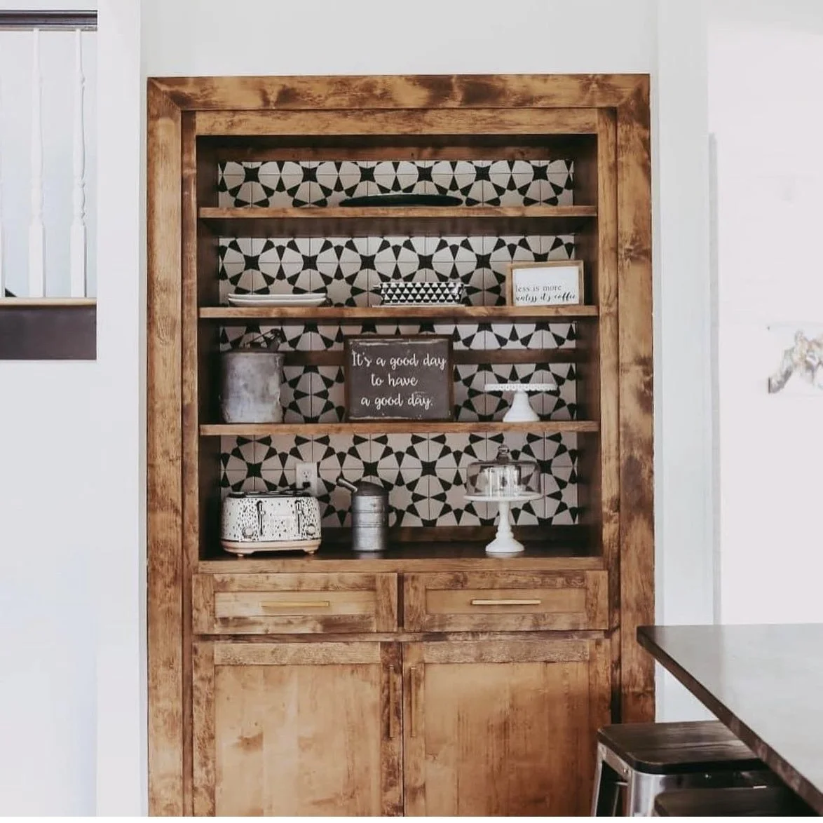 Wooden shelving unit with decorative black and white patterned background, displaying small kitchen decor items including a framed quote, a cake stand with a cake, a vintage toaster, and a small potted plant.