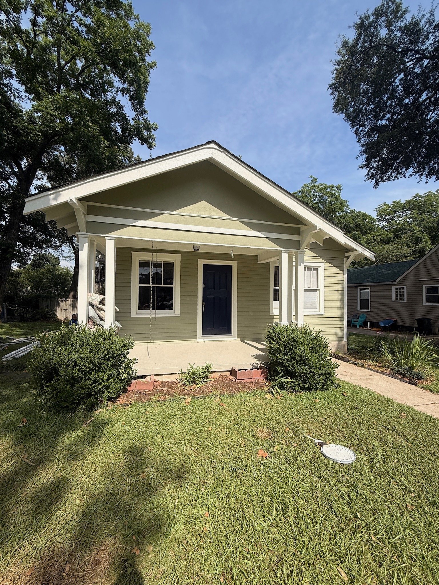A light green house with white trim and a dark blue front door, front porch with bush plants, surrounded by a grassy yard, with trees and a blue sky in the background.