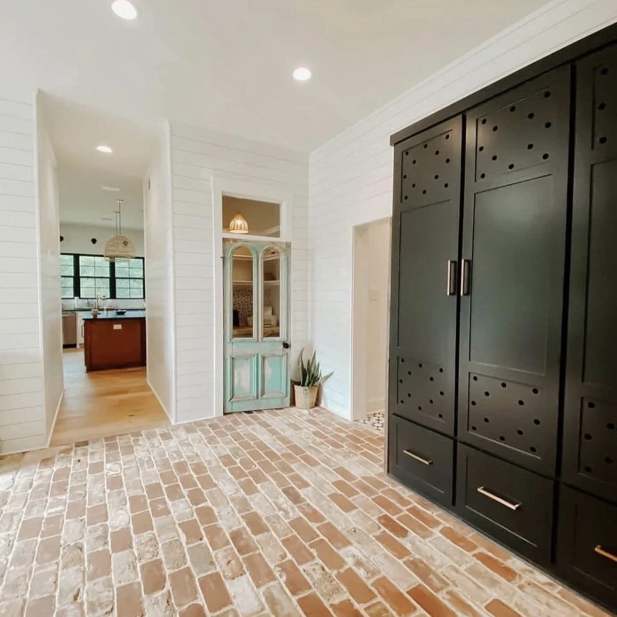 Room with brick floor, white shiplap walls, black storage cabinet, and a vintage door with a plant nearby.