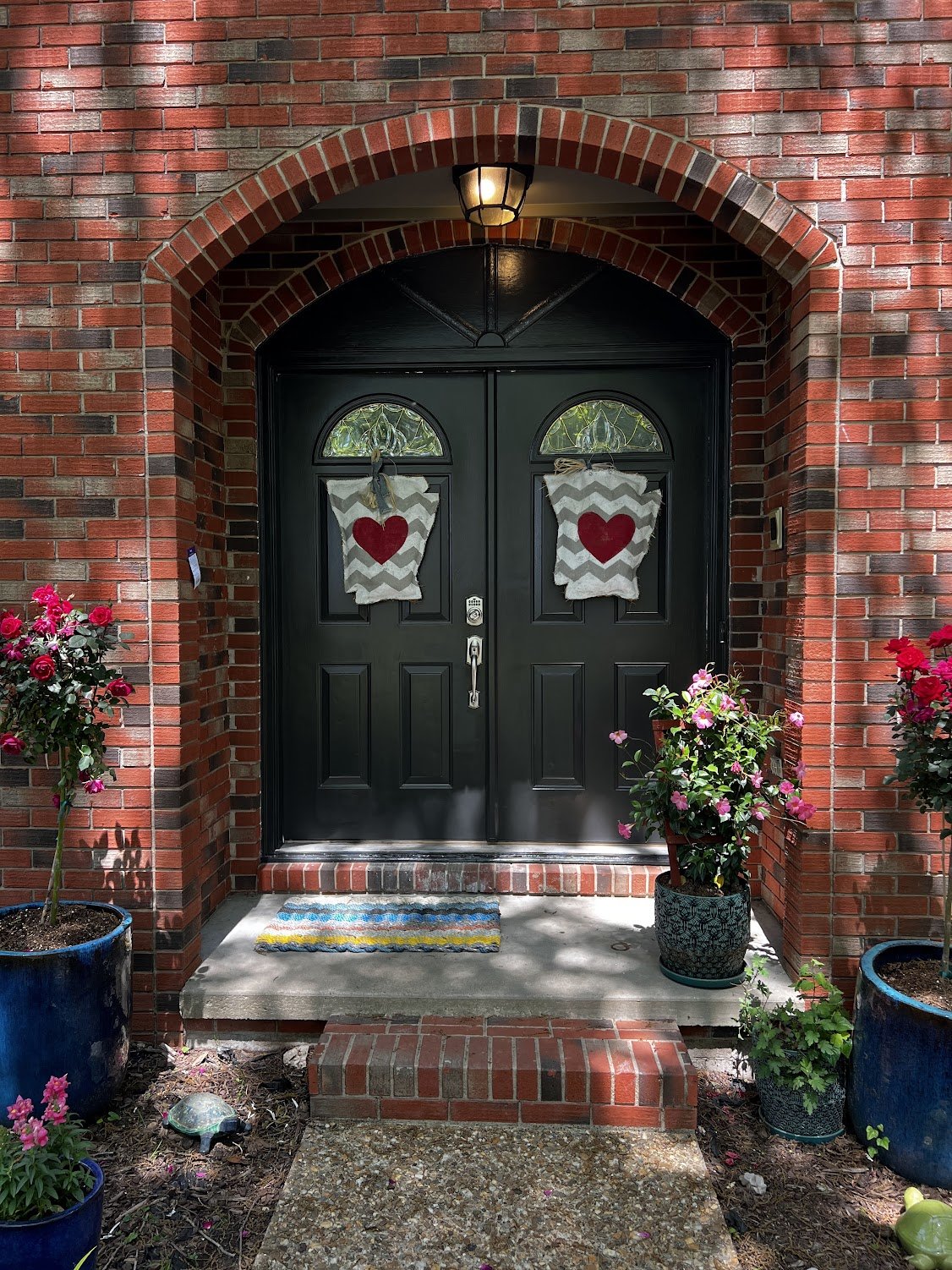 Black front door with two wreaths featuring red hearts, surrounded by potted pink and red flowering plants and a colorful striped doormat, set within a brick porch.