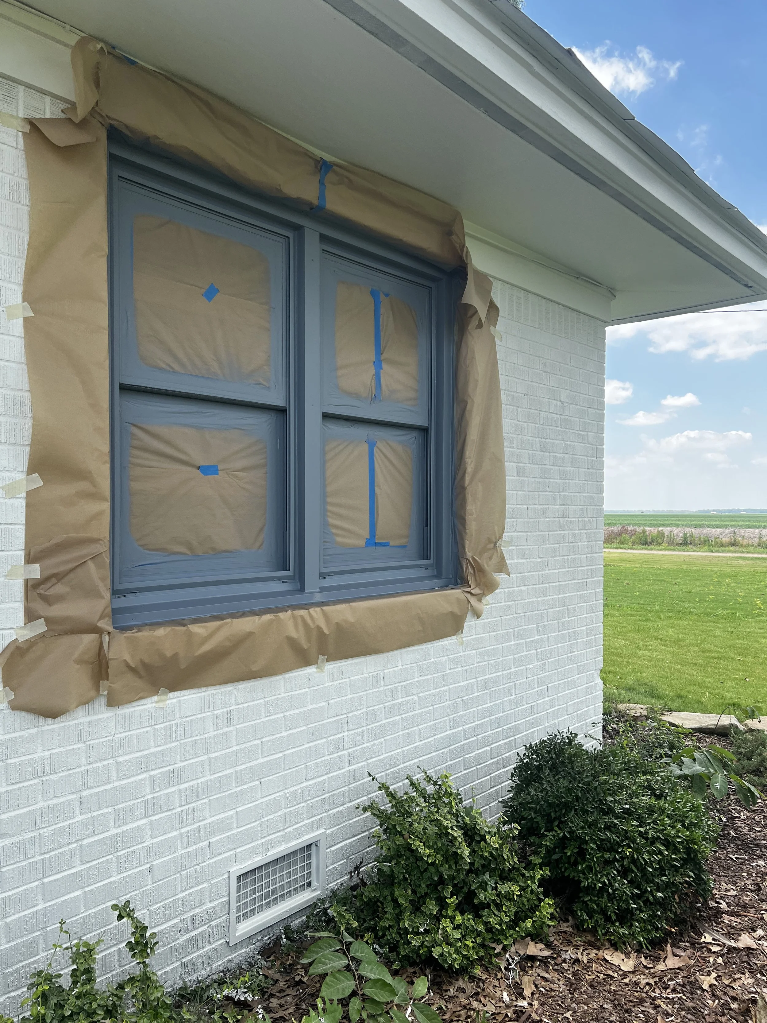 The house window is covered with brown paper and blue painter's tape, indicating it is being prepared for painting or insulation work. The house has white brick siding, and there is a vent near the ground along with some bushes and plants outside.