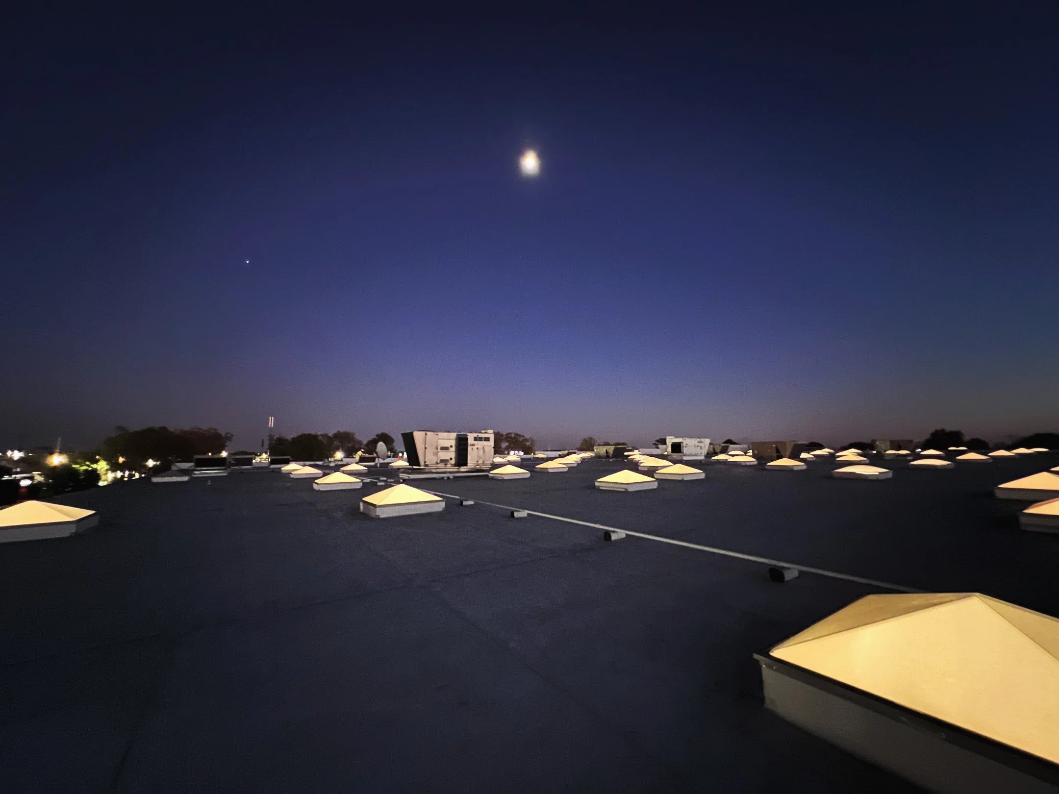 Nighttime rooftop view with several illuminated skylights, dark sky with the moon and stars visible, and trees in the distance.