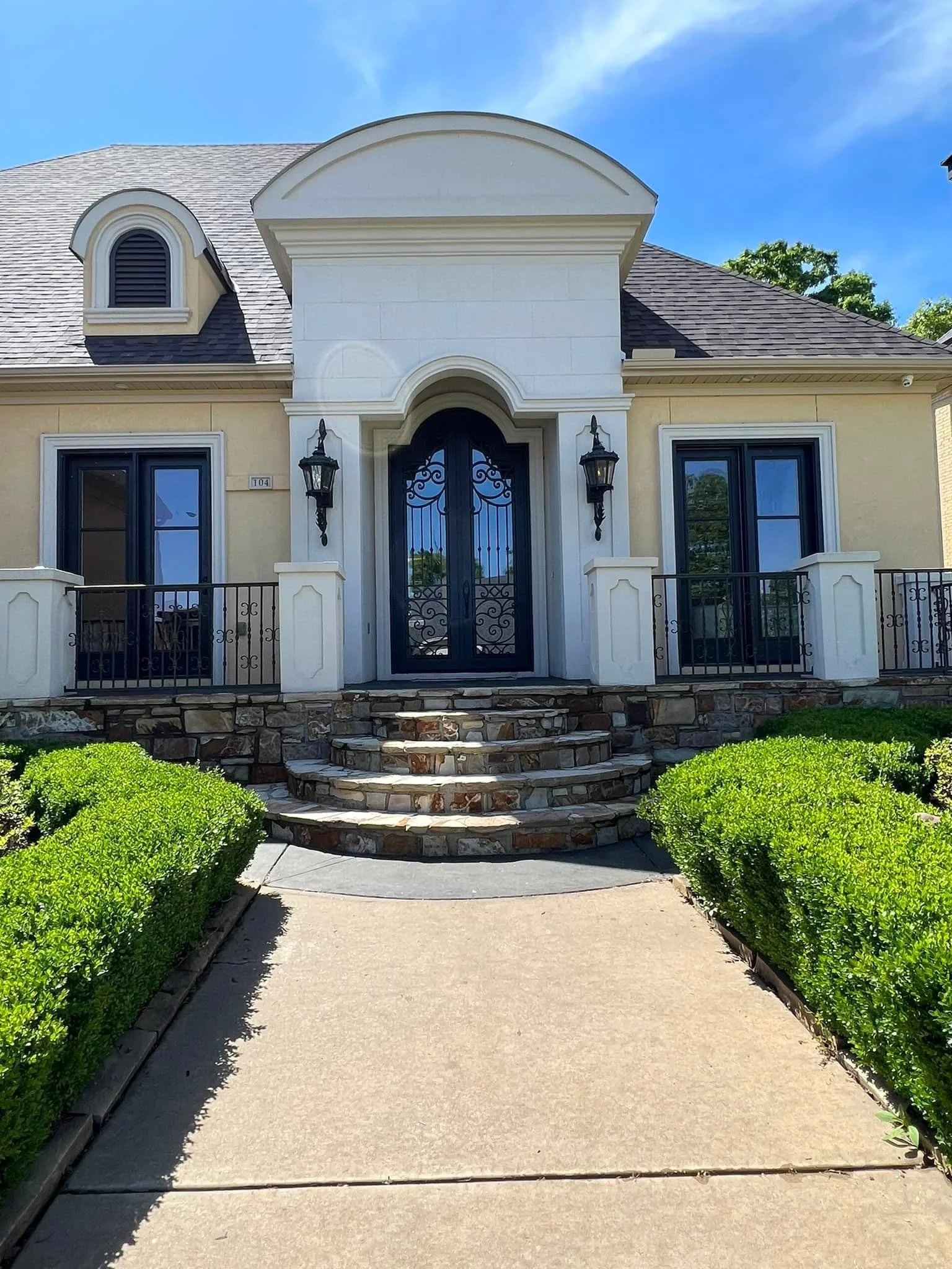 Front view of a house with stone steps leading to a black wrought iron door, flanked by two black wall lanterns, with green bushes lining the walkway, and a clear blue sky overhead.