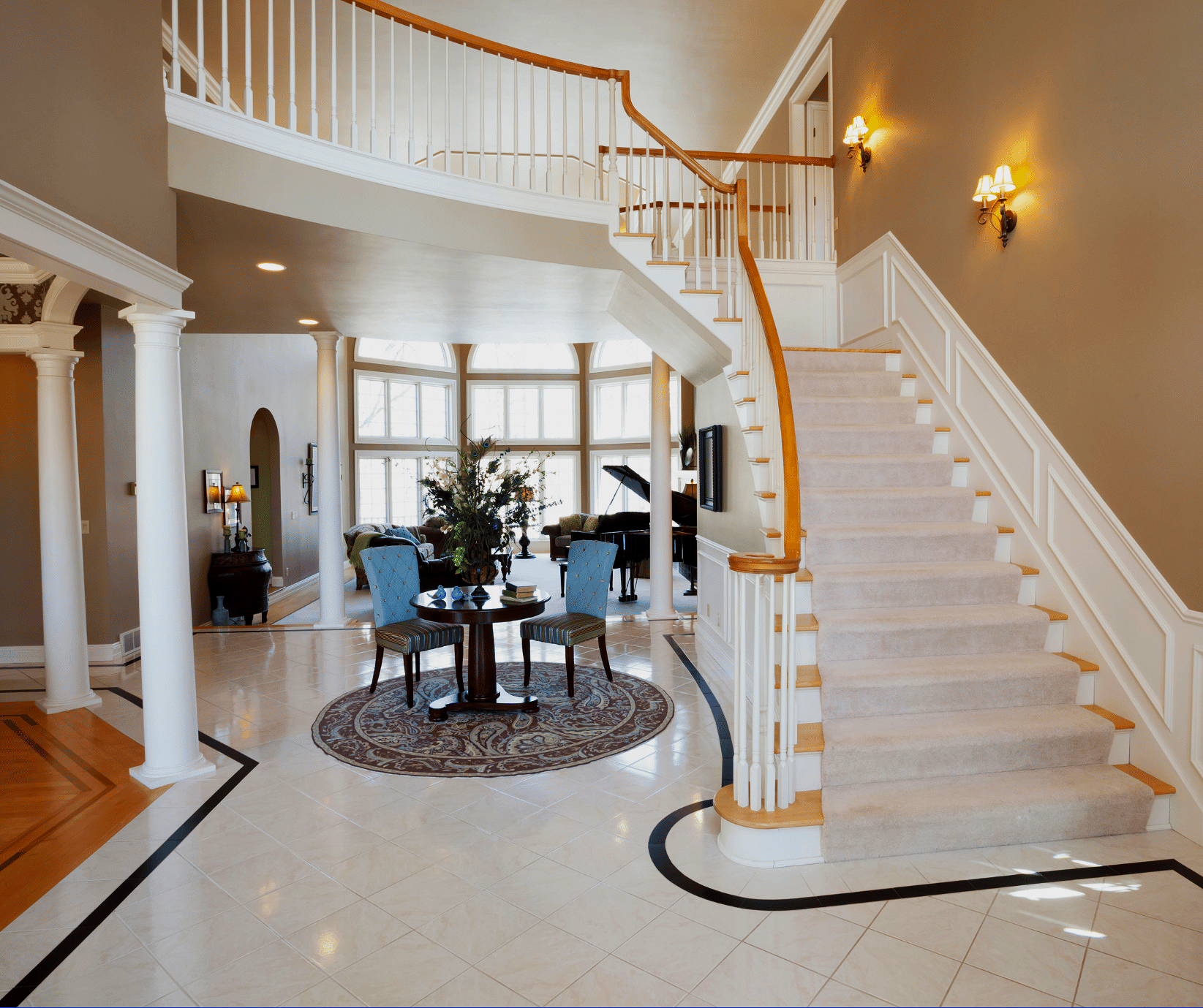 Interior of a spacious, well-lit home with a curved staircase with a wooden handrail and carpeted steps, a round dining table with three chairs, large windows, and a living area with a grand piano and seating.