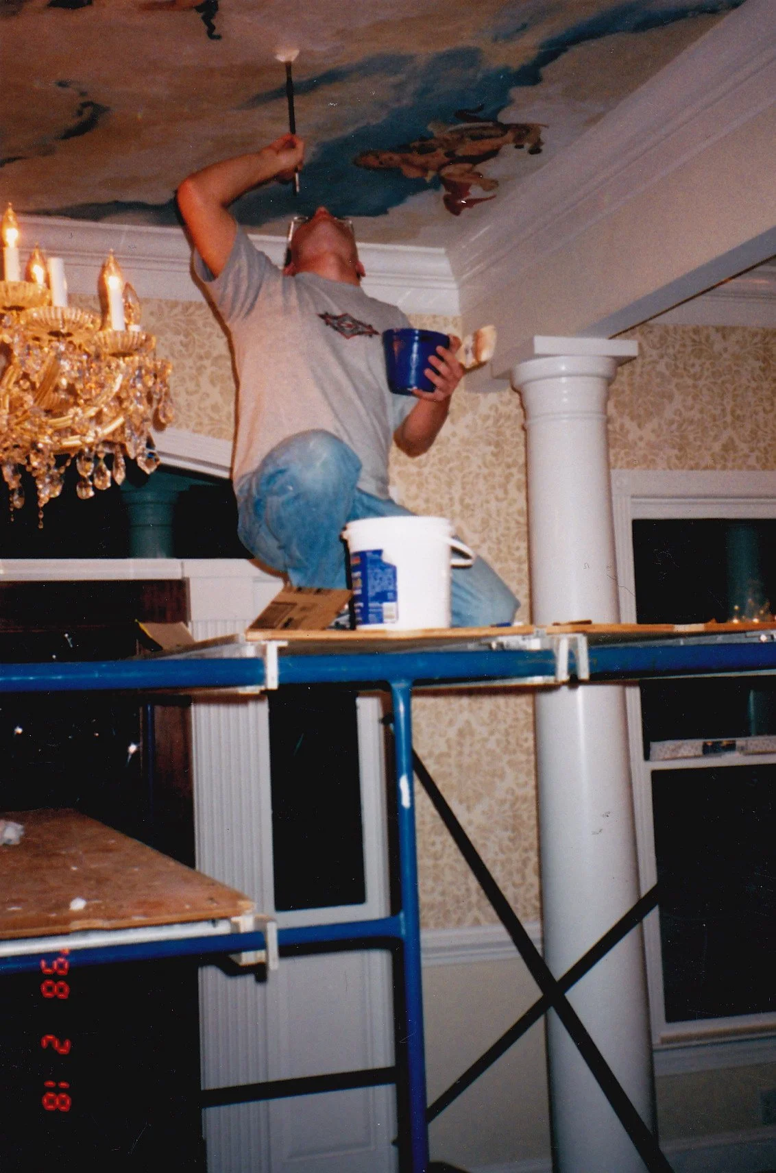 Person painting a ceiling with a small brush while kneeling on a platform, holding a cup, surrounded by painting supplies and interior decor.