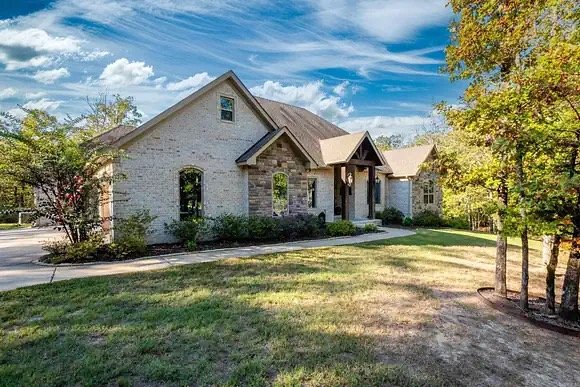 A modern house with a stone facade, front porch, and a lawn, surrounded by trees under a partly cloudy sky.