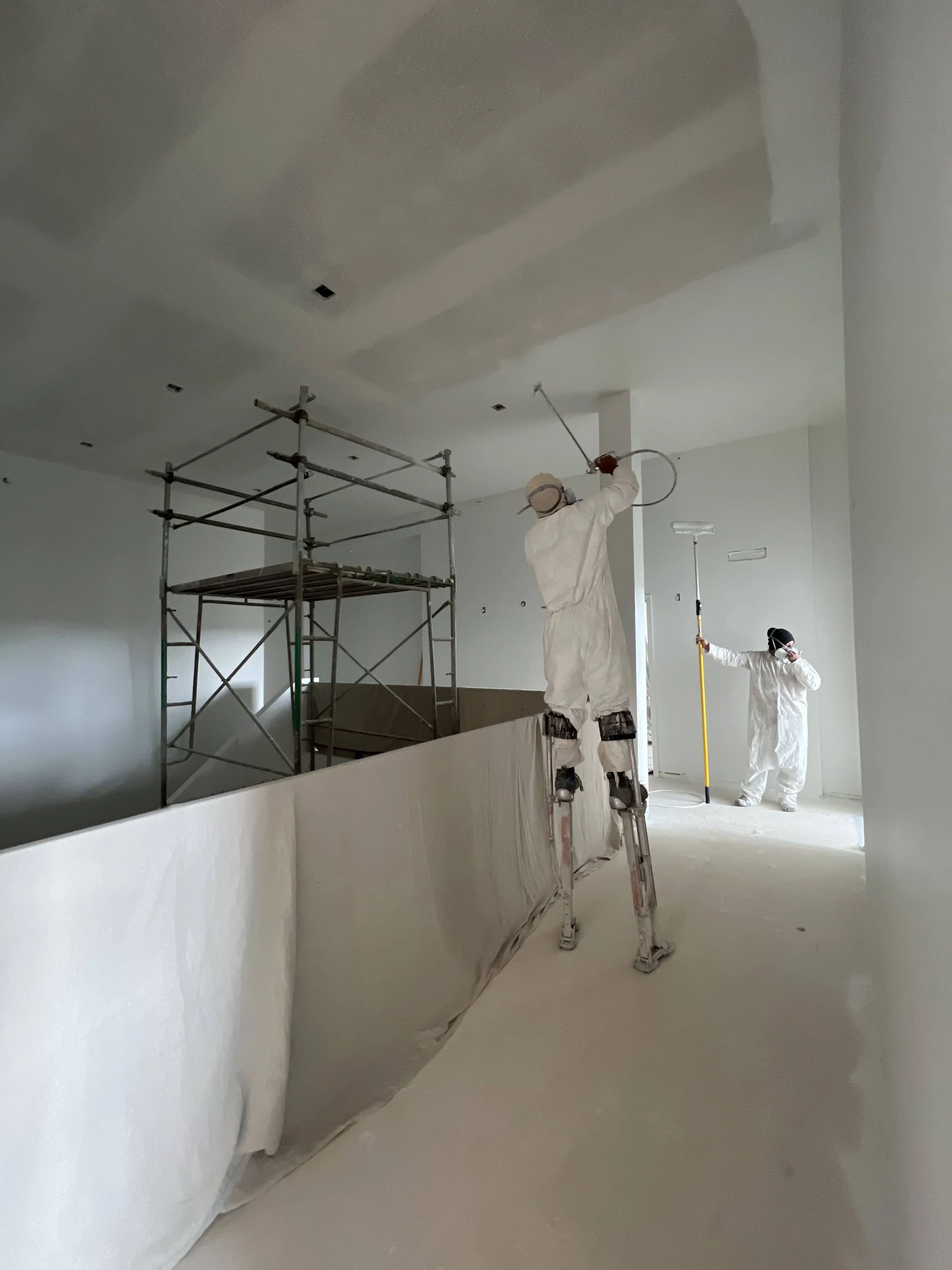 Two workers dressed in white protective suits working on the ceiling of an indoor construction site. One worker is on a ladder using a tool, while the other is on the ground holding a long pole, with scaffolding visible in the background.