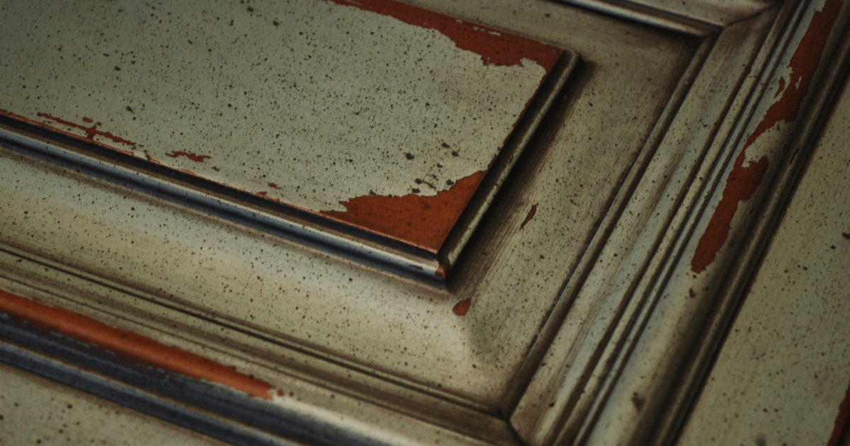 Close-up of a weathered, distressed wooden cabinet door with chipped paint in beige and rust colors.