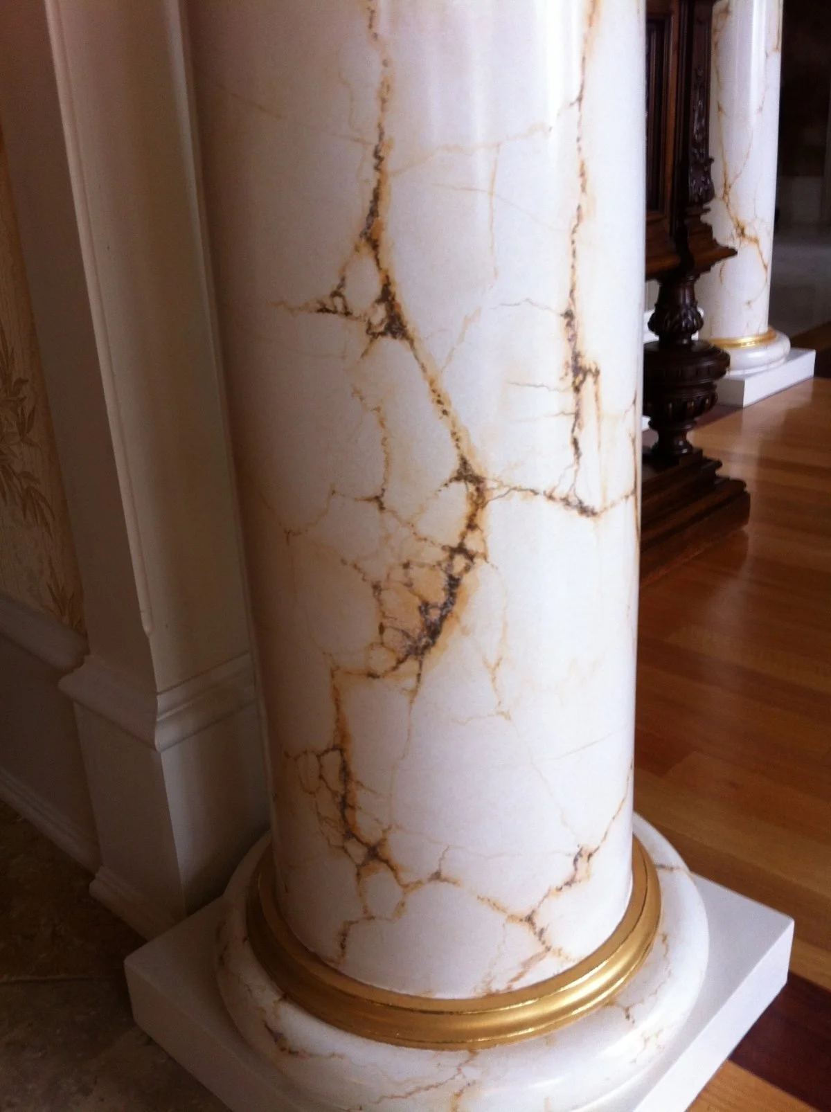 Close-up of a large, decorative marble column with a white base color and brownish-golden veining, situated in an indoor setting with polished hardwood flooring.