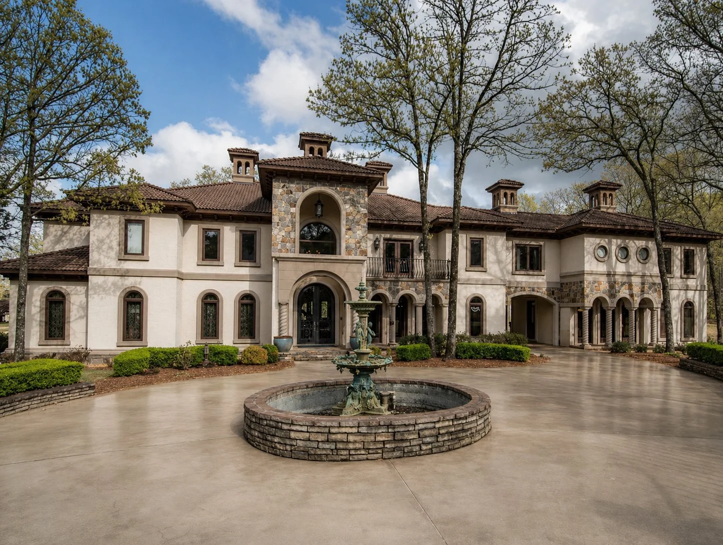 A large mansion with a stone and stucco exterior, arched windows, and a fountain in front, surrounded by trees and a concrete driveway.
