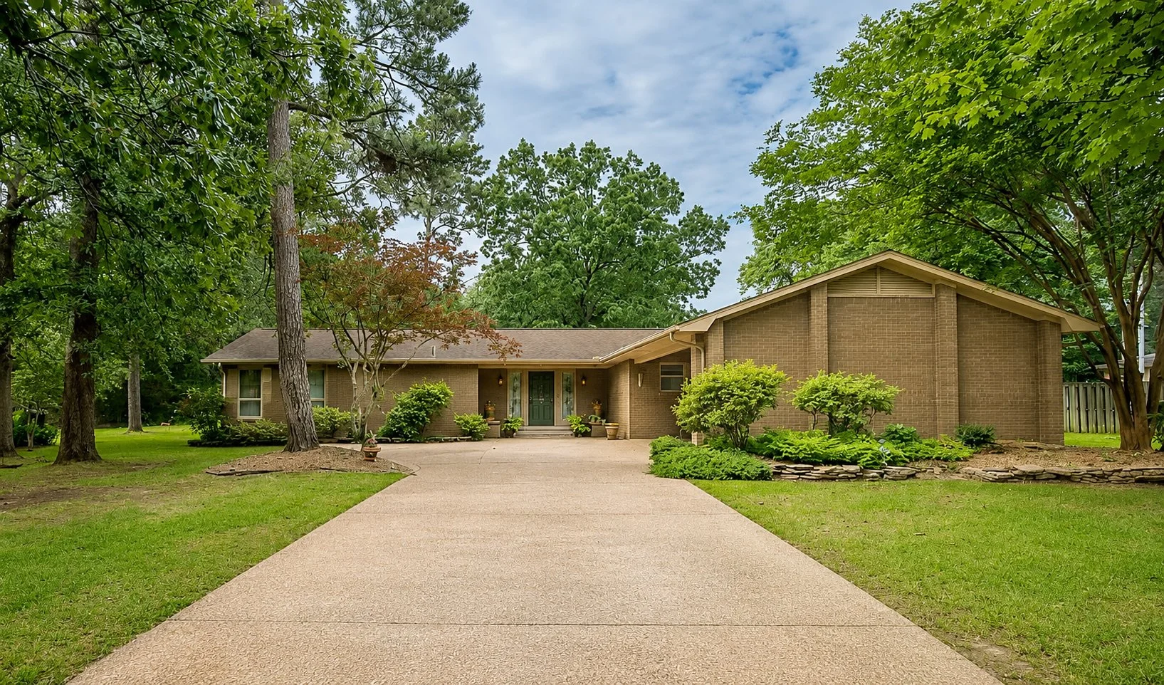 Single-story brick house with a green front door, surrounded by green trees and shrubs, and a concrete driveway leading to the entrance.