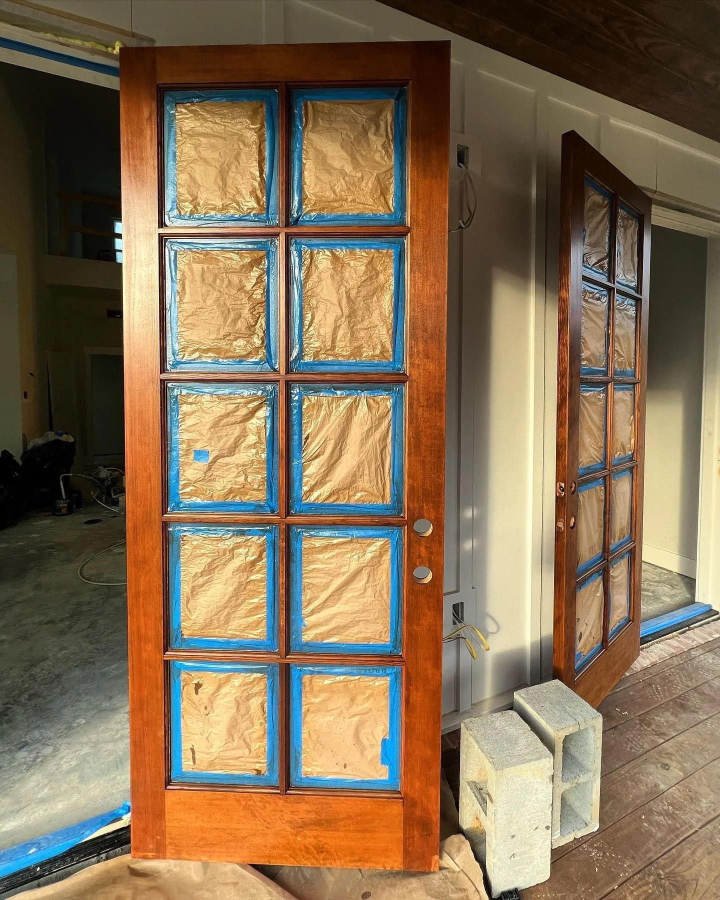 A wooden door with its glass panels covered with brown paper and blue painter's tape, undergoing renovation or painting process in a house under construction.