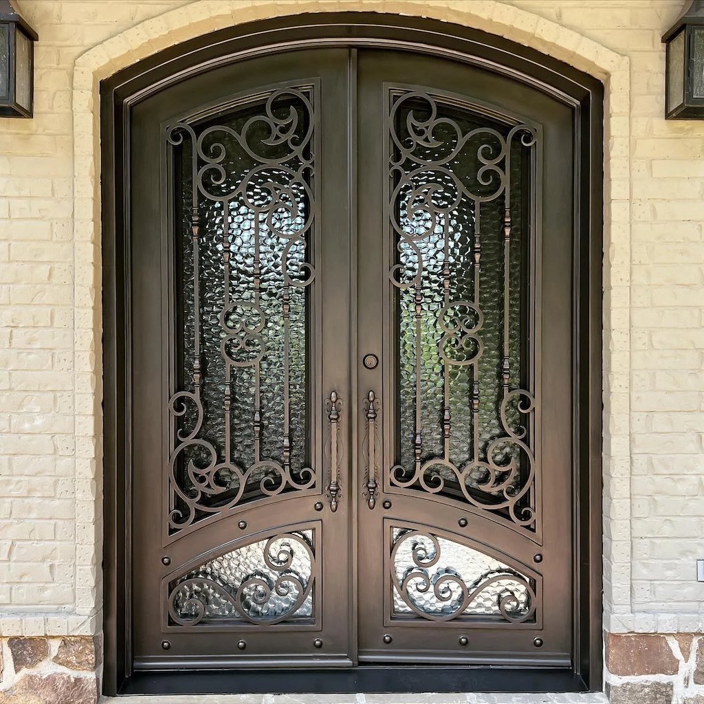 Decorative metal double door with intricate scrollwork design, set in a brick exterior wall with stone accents at the base.