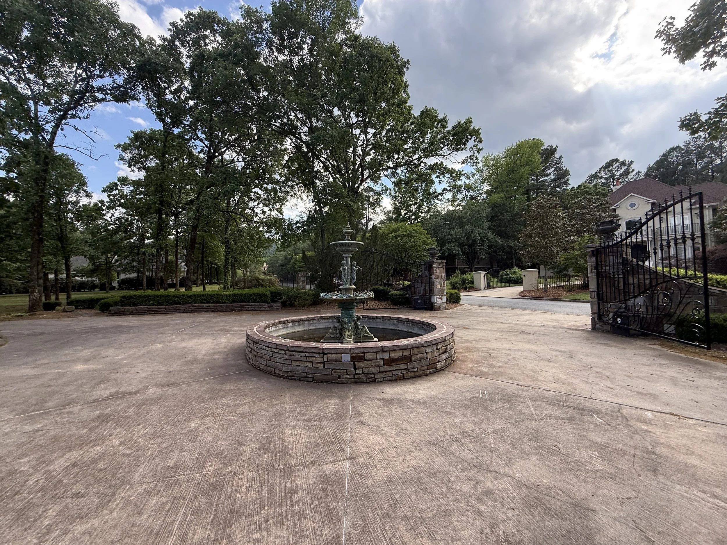 A large round brick fountain with multiple tiers and decorative sculptures stands in a spacious paved courtyard surrounded by tall trees and lush greenery. A black wrought iron gate and a white house with a porch can be seen in the background under a partly cloudy sky.