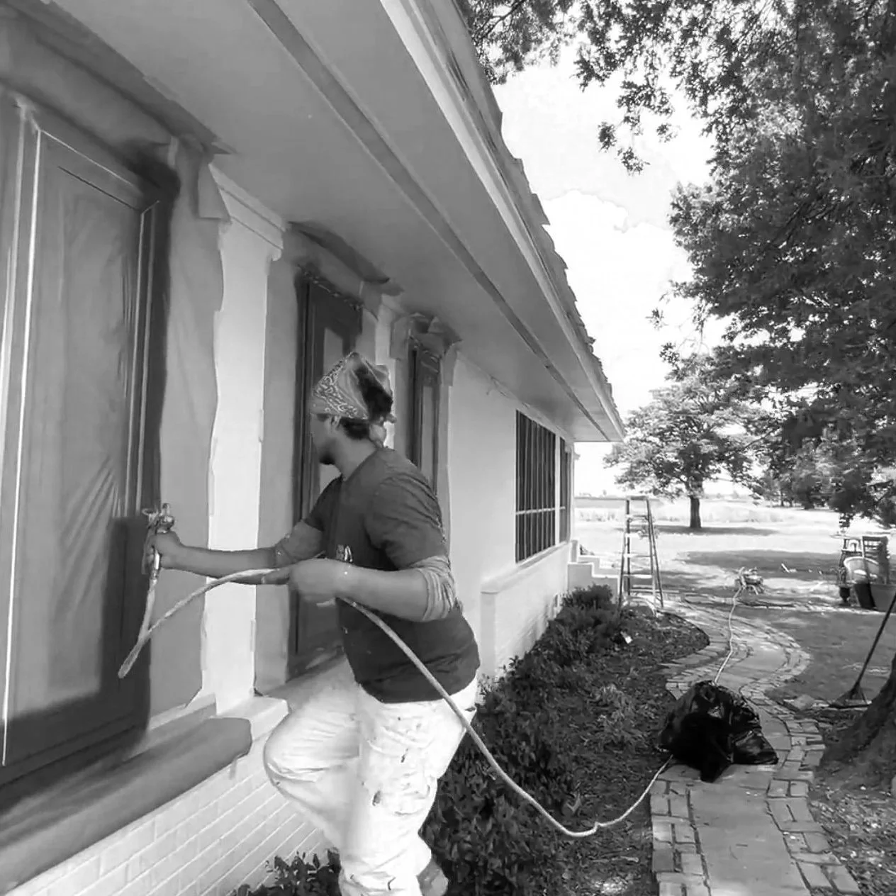 A person wearing a red bandana and gray shirt is power washing the exterior window frames of a house with a garden hose, while standing on a brick pathway next to shrubbery and a grassy yard with trees.