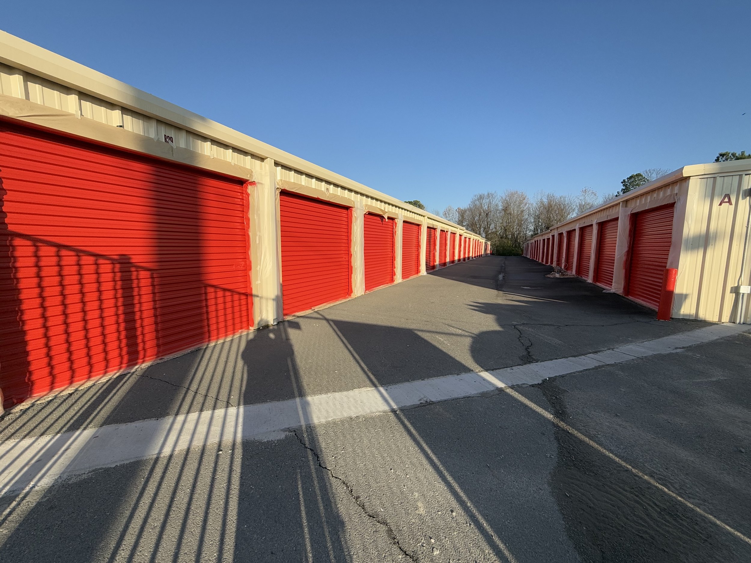 A row of storage units with red roll-up doors and beige walls, casting long shadows on the asphalt ground under a clear blue sky.
