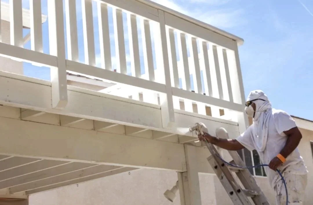 A person in white protective gear, face mask, and sunglasses power washing a white railing on a balcony outside a building.