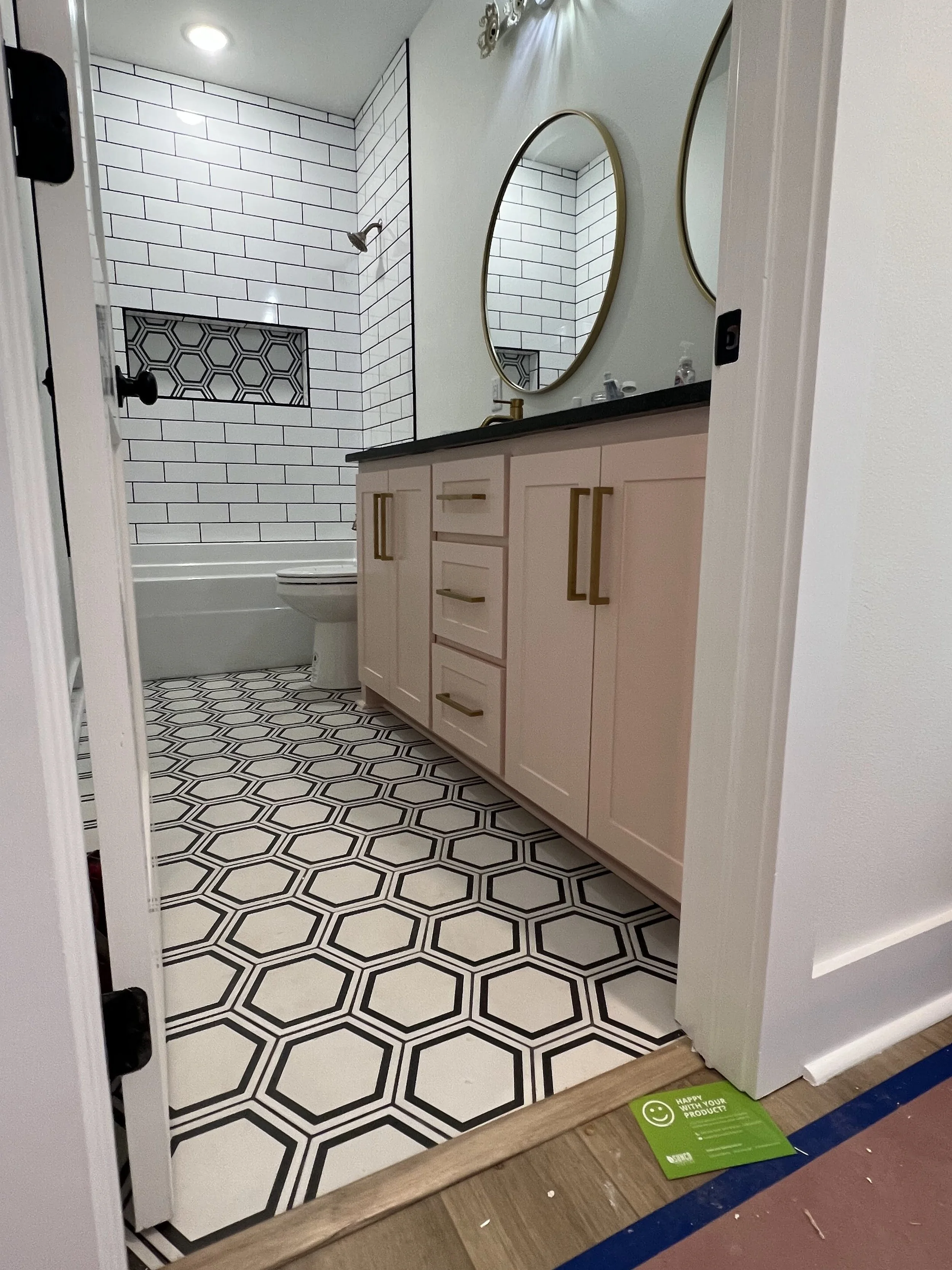 A bathroom with a white vanity featuring gold handles, a black countertop, and two oval mirrors above it. The shower area has white subway tiles with a geometric black and white accent tile in the niche. The floor has black and white hexagonal tiles.