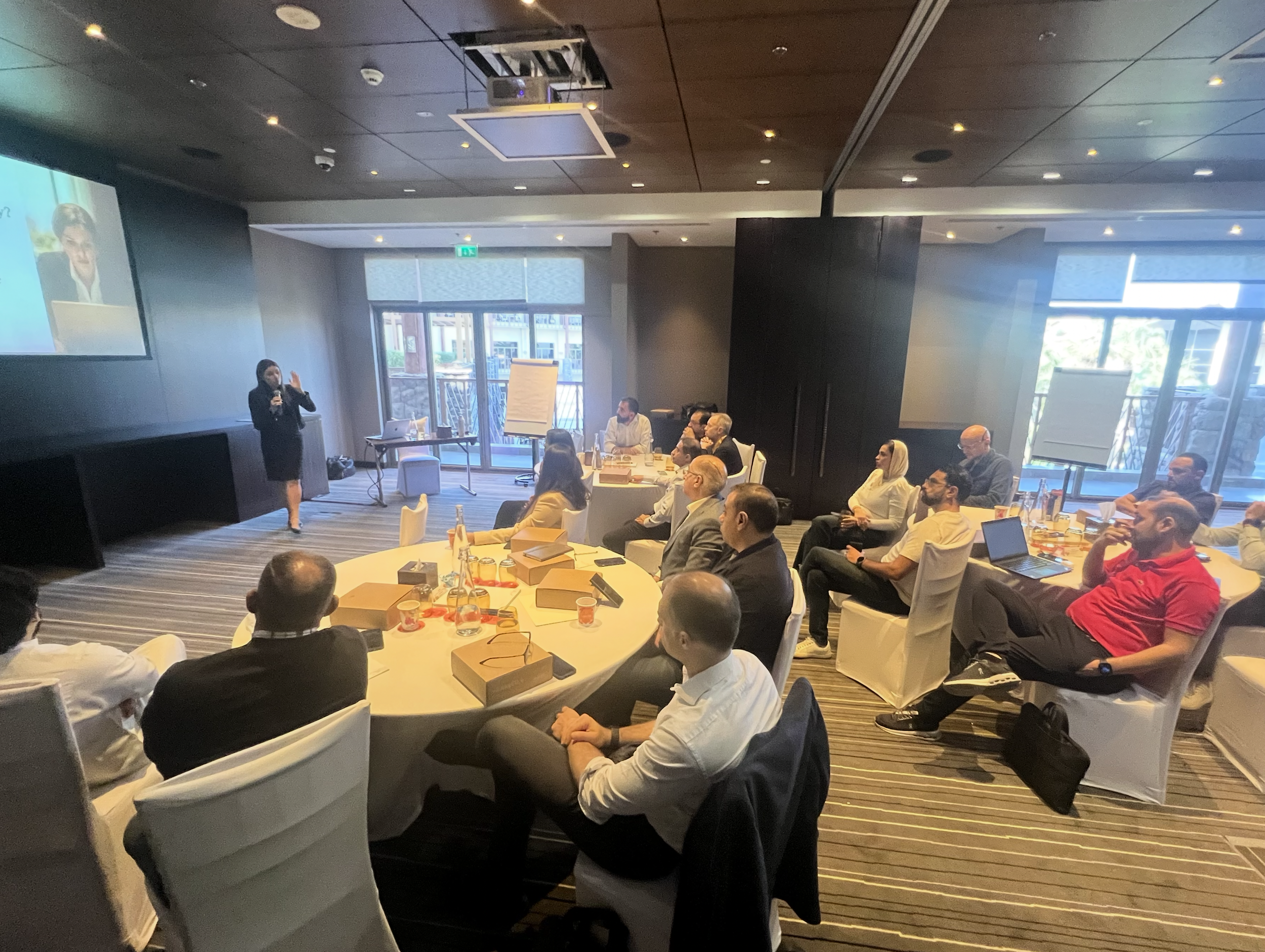 Portrait of a woman with dark hair styled in an updo, smiling, wearing a white blazer, against a plain gray background.