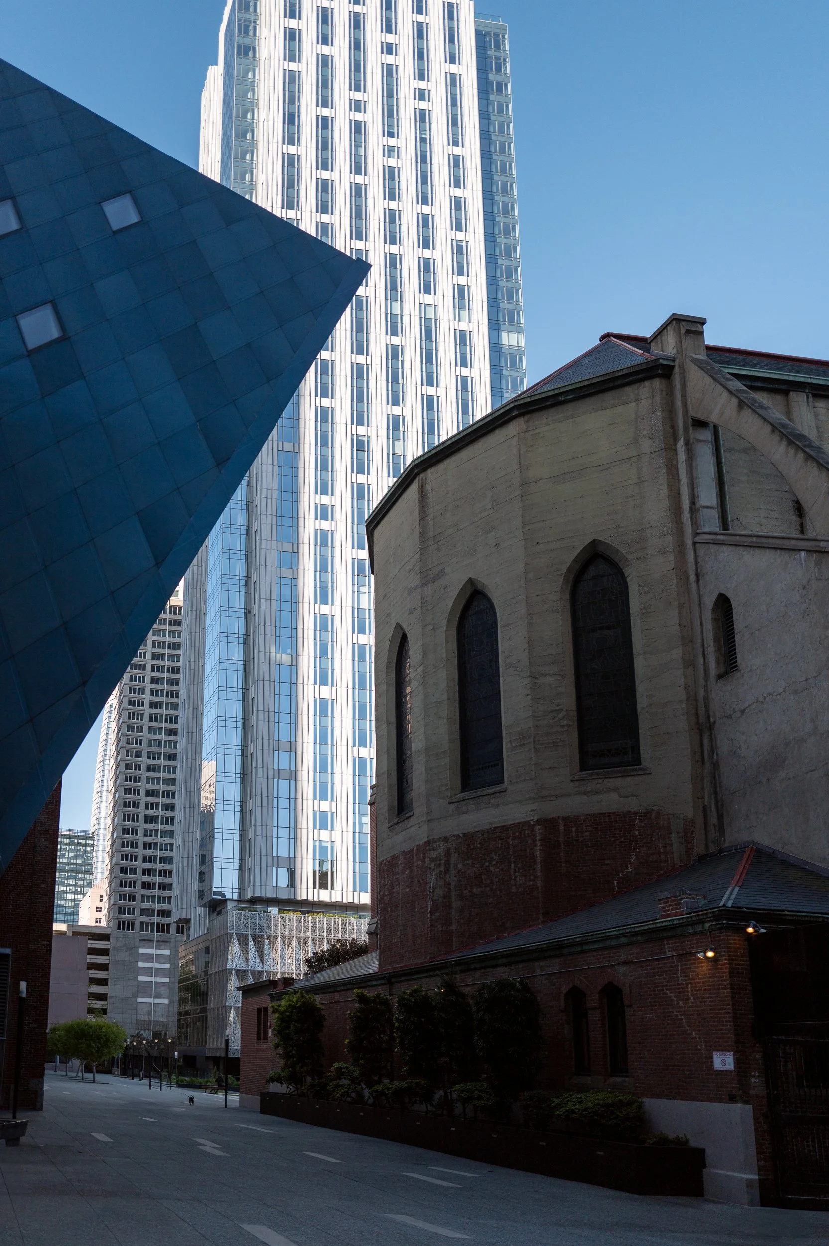 Jeunes arbres devant un bâtiment ancien en pierre, entouré de gratte-ciel modernes en verre, dans une rue urbaine moderne.