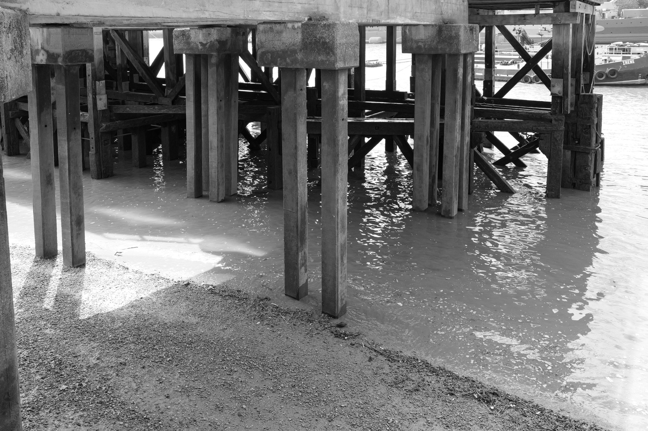 Structure en bois sous un pont au bord de l'eau, vue en noir et blanc.