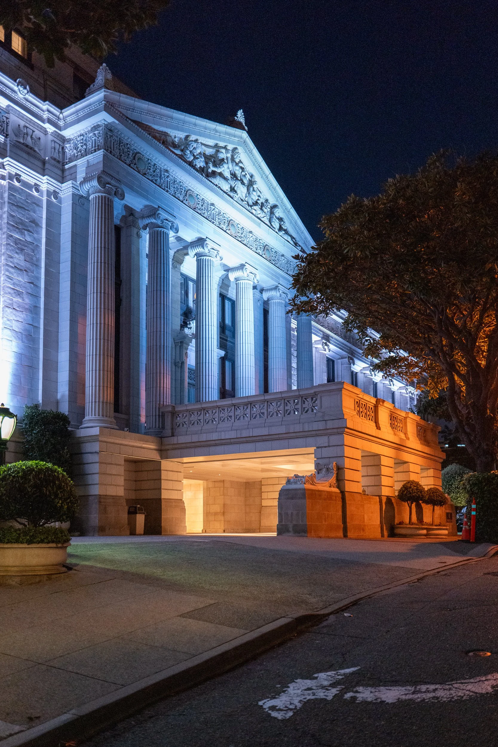 Bâtiment historique éclairé la nuit, avec des colonnes classiques et des détails architecturaux ornés, entouré d'arbres.