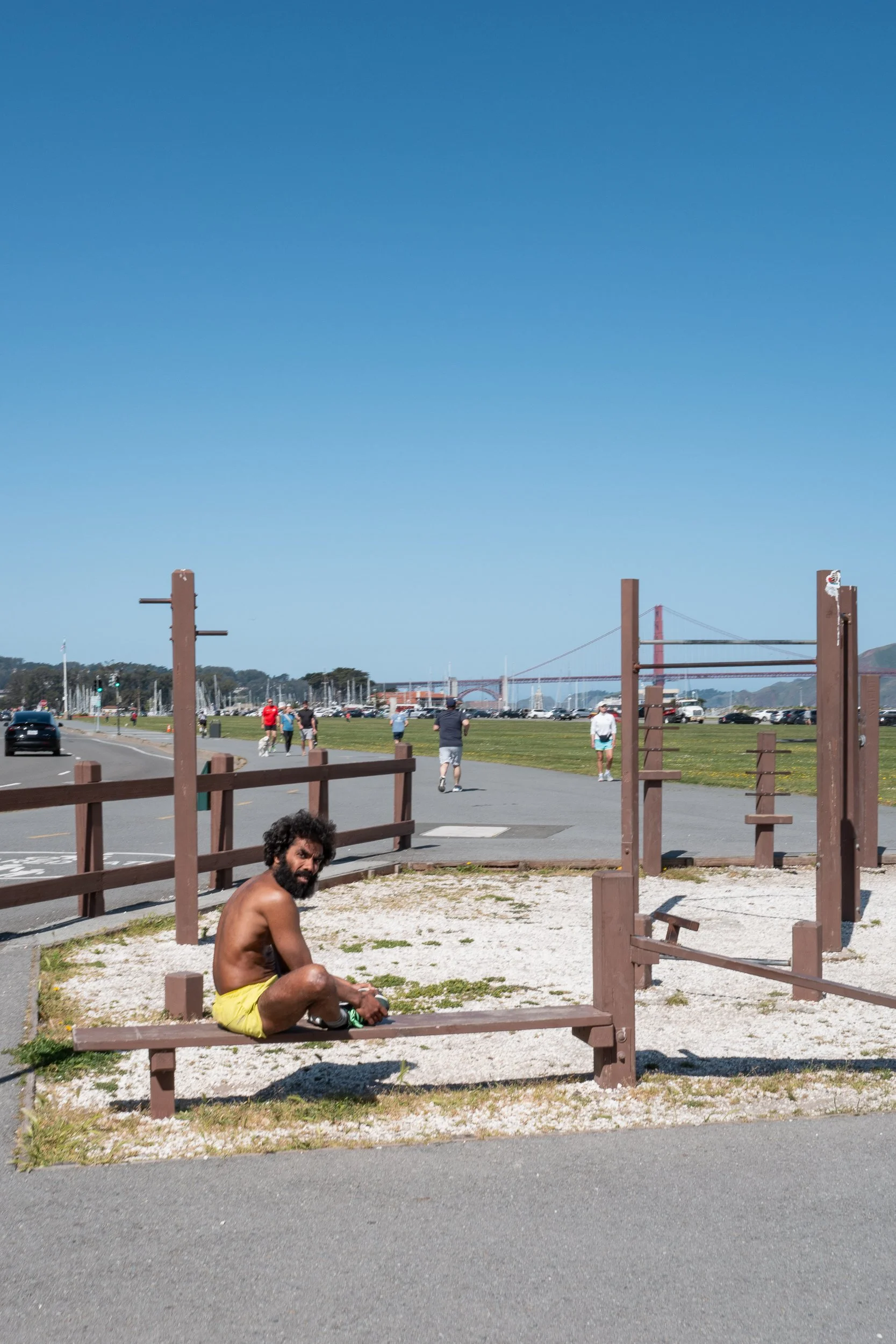 Un homme torse nu, assis sur un banc dans un espace de sport en plein air, avec la baie de San Francisco et le pont du Golden Gate en arrière-plan, par une journée ensoleillée.