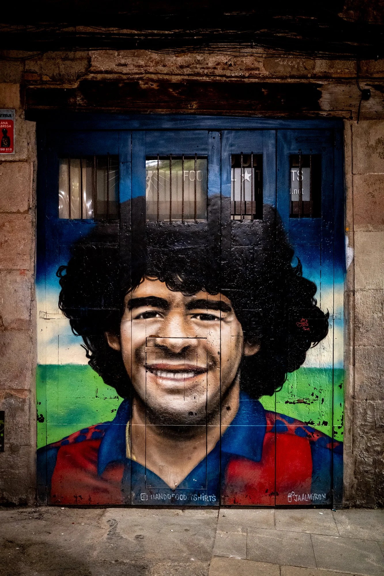 Une fresque murale d'un homme souriant avec des cheveux bouclés, portant une chemise rouge et bleue, sur un fond de paysage vert et bleu.