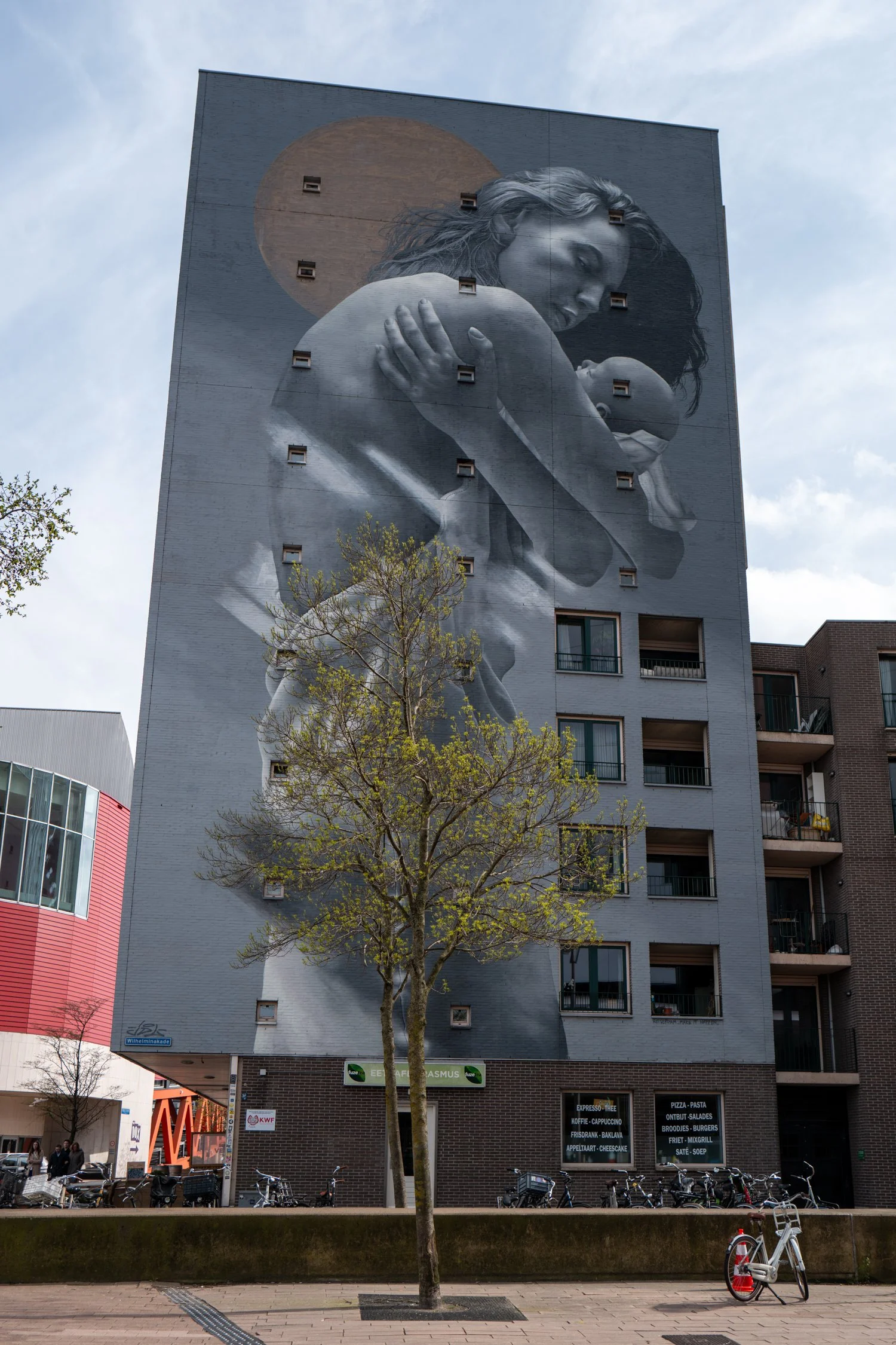 Une fresque murale en noir et blanc représentant une femme avec un bébé, avec un fond représentant la lune ou un soleil, située sur un bâtiment d'appartements en ville. Il y a des arbres, des vélos et des commerces au rez-de-chaussée.